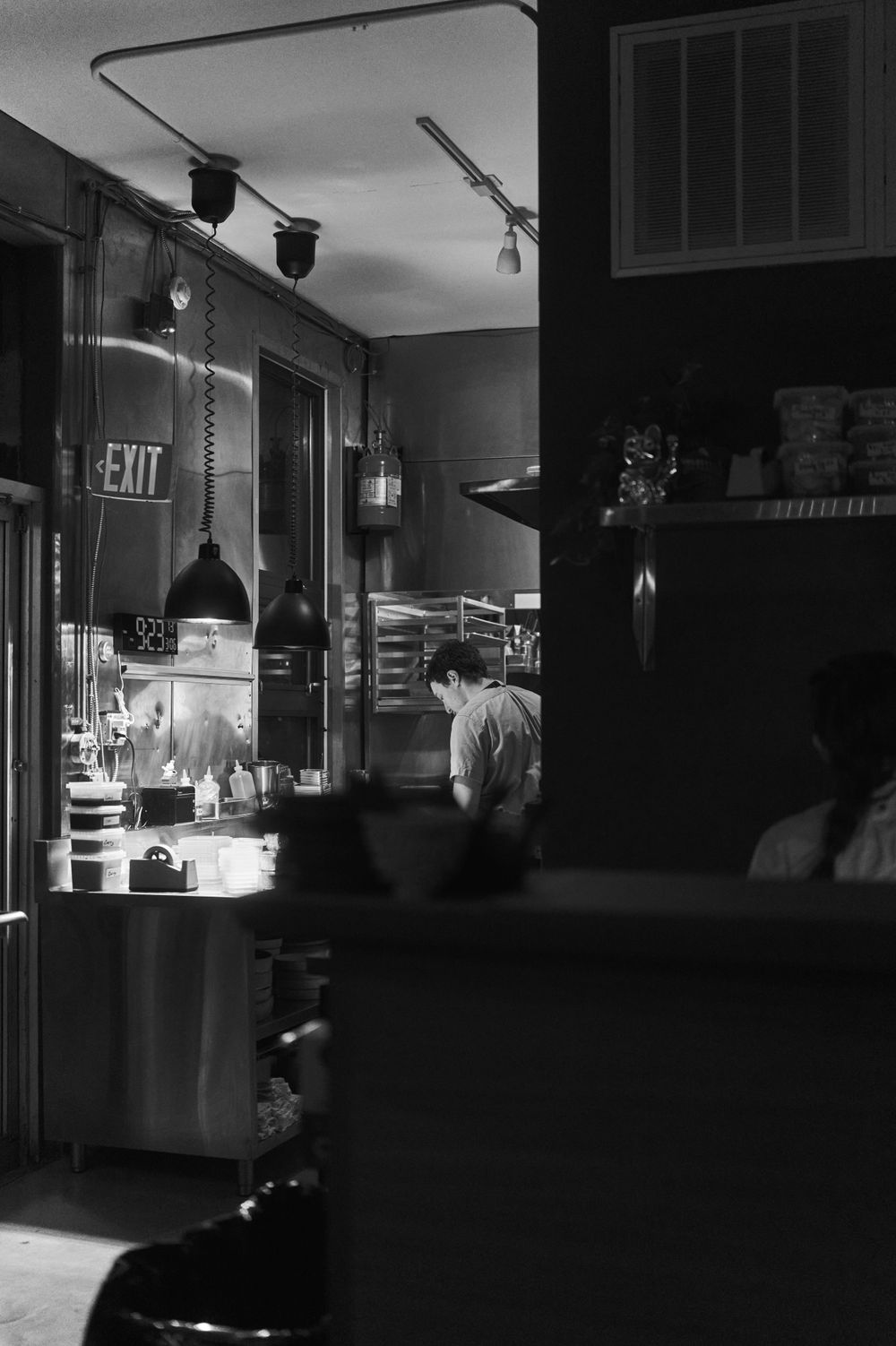 Moody black and white view of a restaurant kitchen with exit sign and overhead lighting.