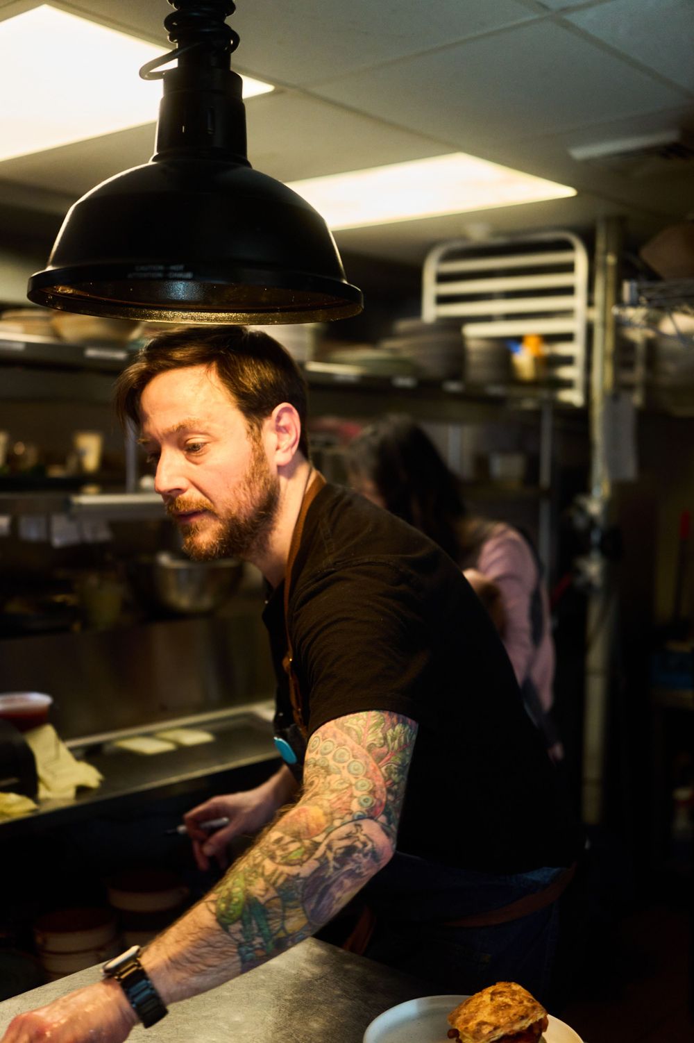 Kitchen staff member with tattoos working under industrial pendant lamp in restaurant kitchen.