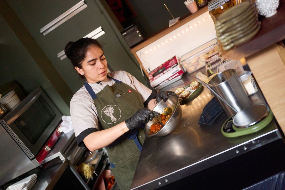 Restaurant worker in green apron preparing food at stainless steel prep station in commercial kitchen.