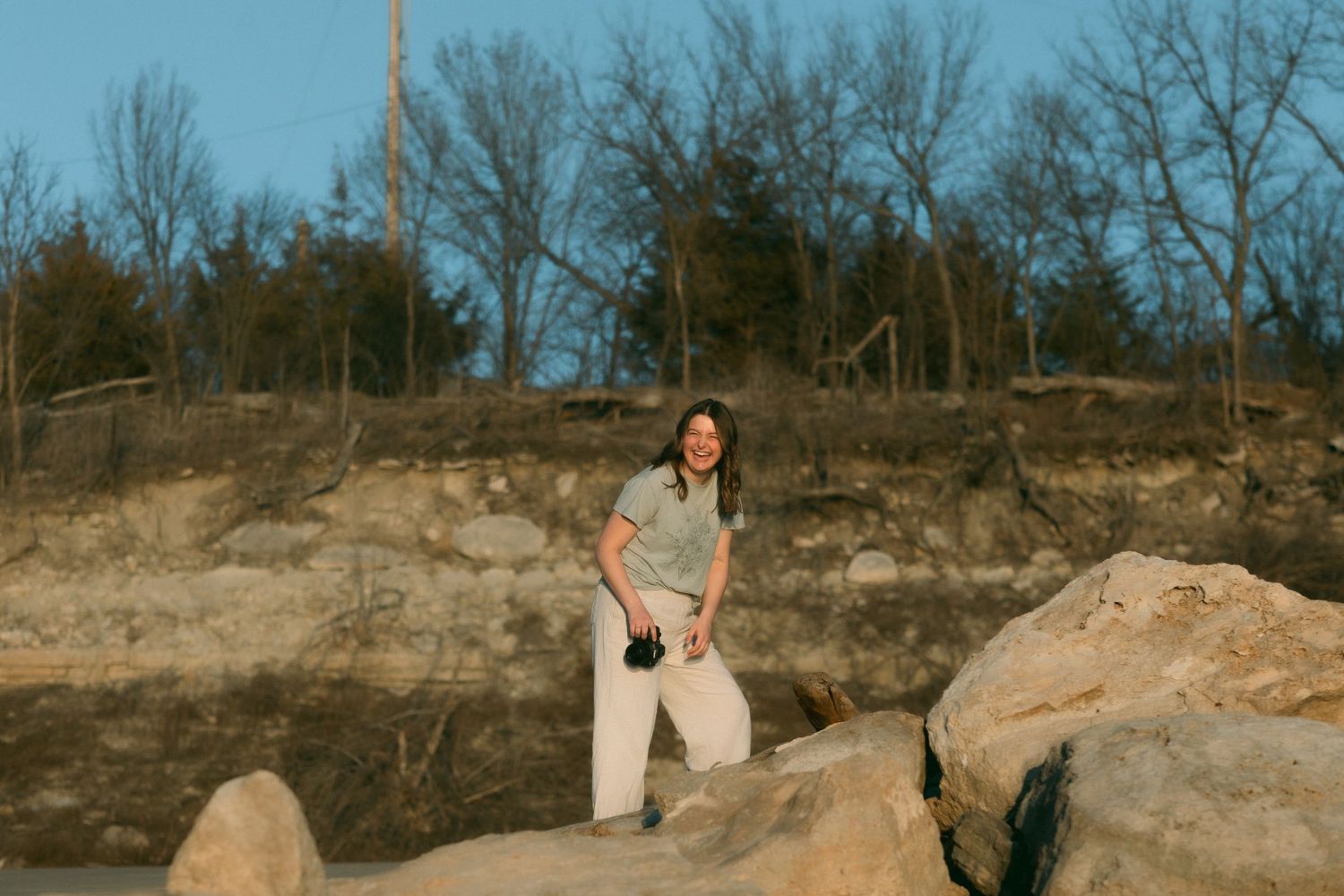 Emily Mae laughing while climbing a rock. She is holding her Canon R6 Mark II in one hand and smiling downwards
