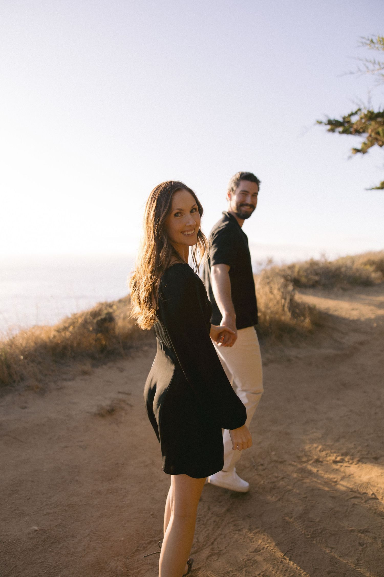Couple standing on hilltop trail at sunset with scenic mountain views in background.