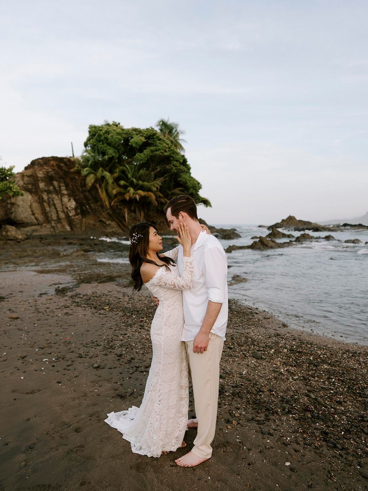 Couple in white attire walking along rocky tropical beach at sunset.