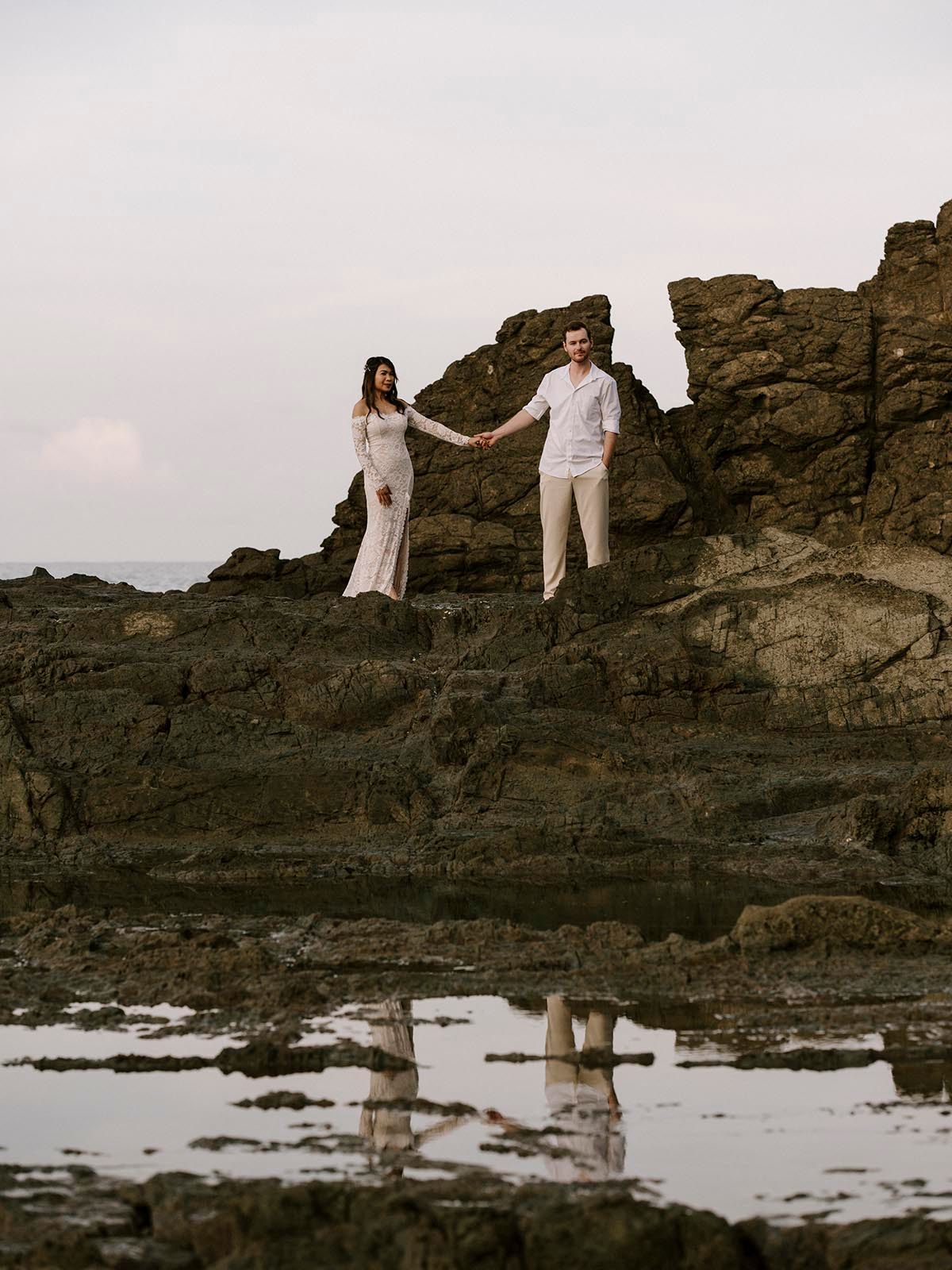 Two people in white clothing reflected in tidal pools on volcanic rocks at sunset.