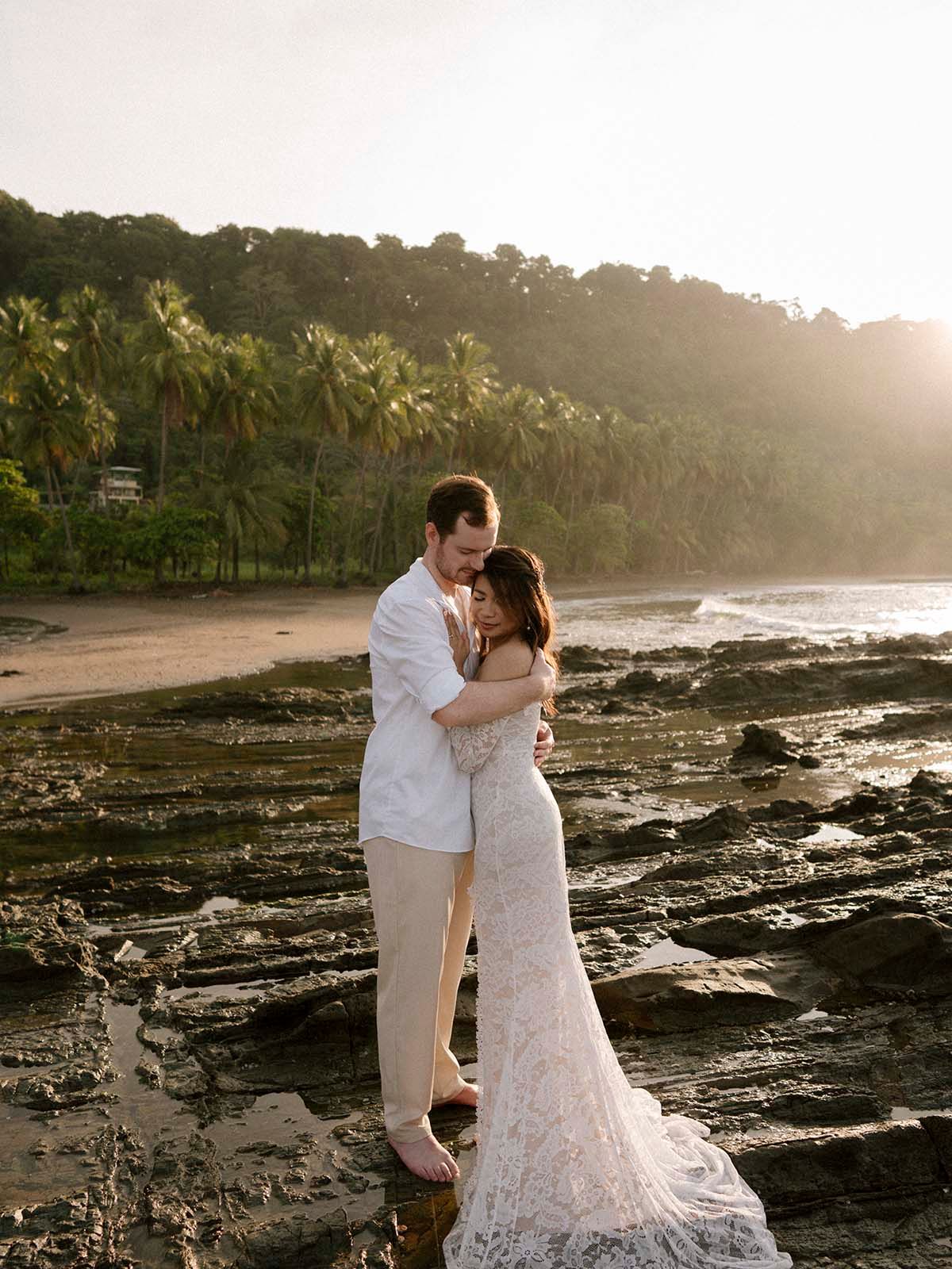 Wedding couple embracing on volcanic rocks with palm trees and ocean in background.