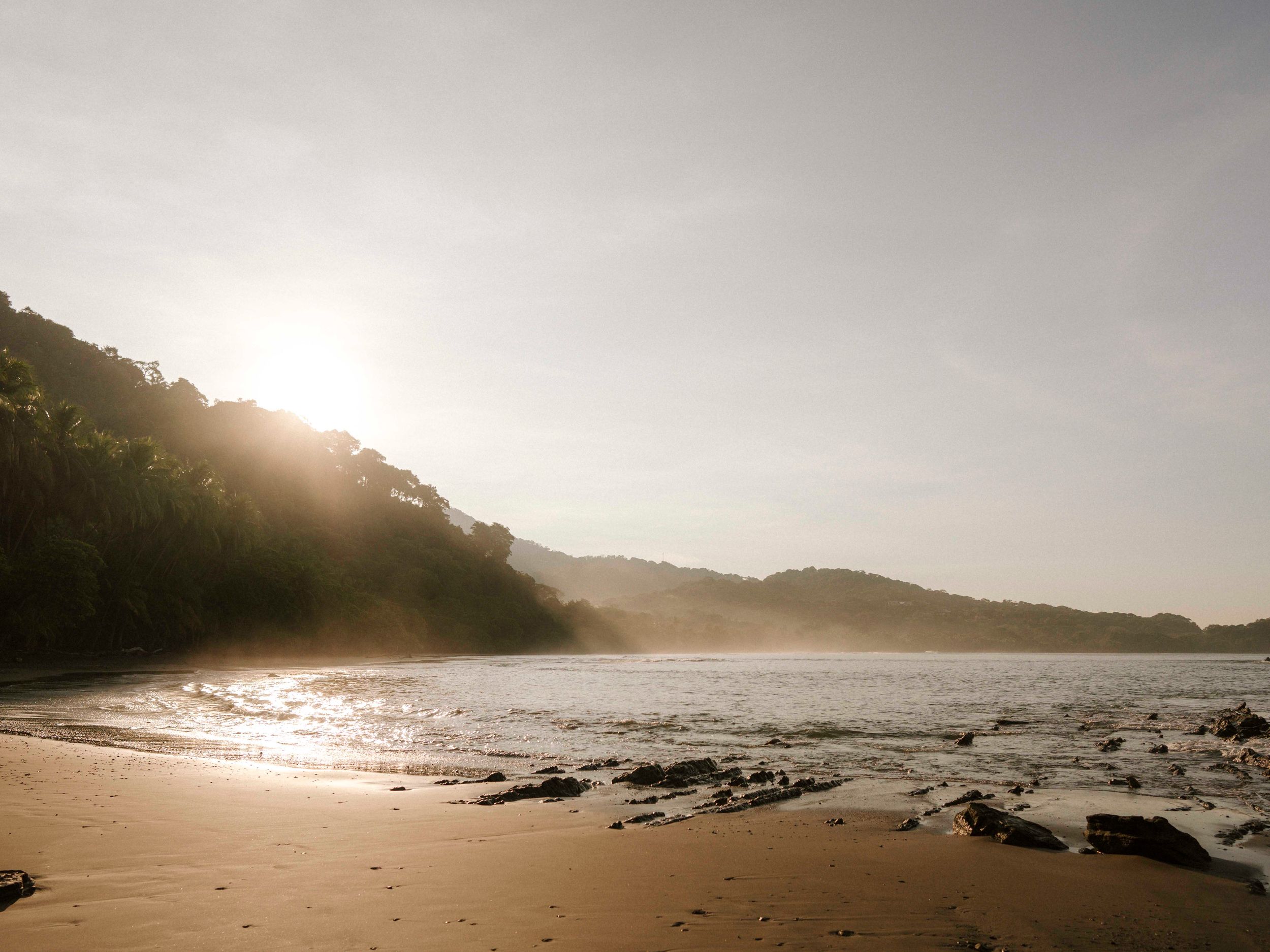 Misty beach shoreline with rocky outcrops and mountains in the distance during golden hour.