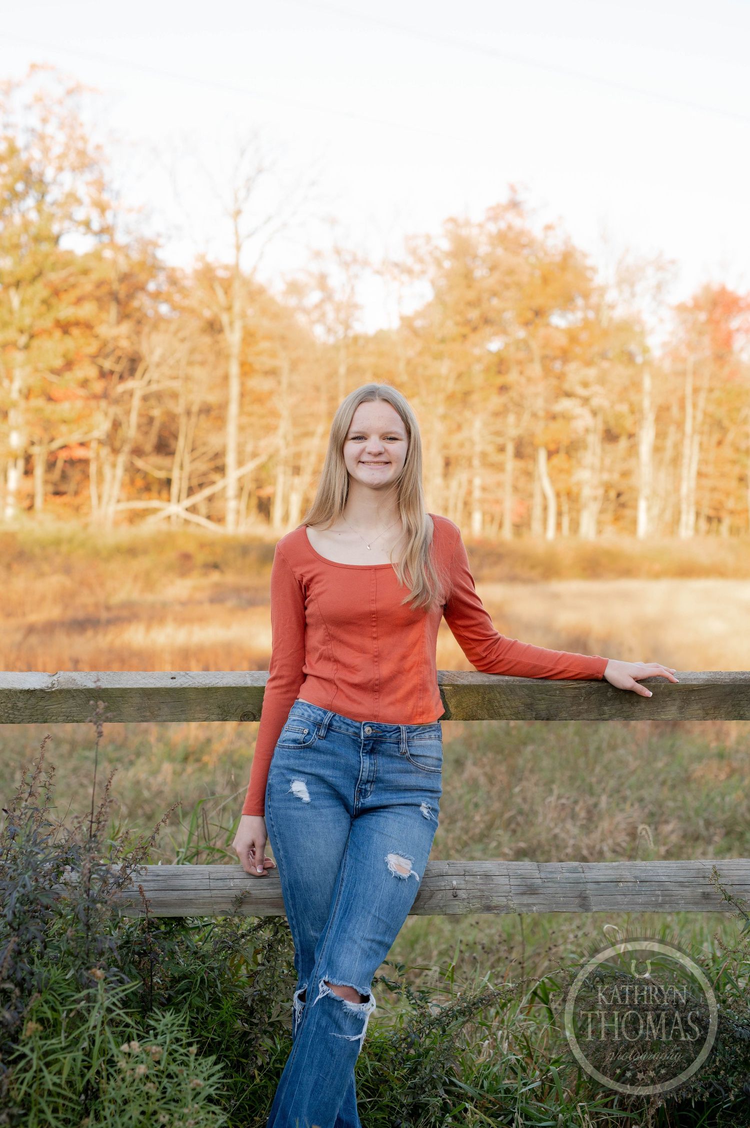 Casual autumn portrait against wooden fence with distressed jeans and orange top in golden light.