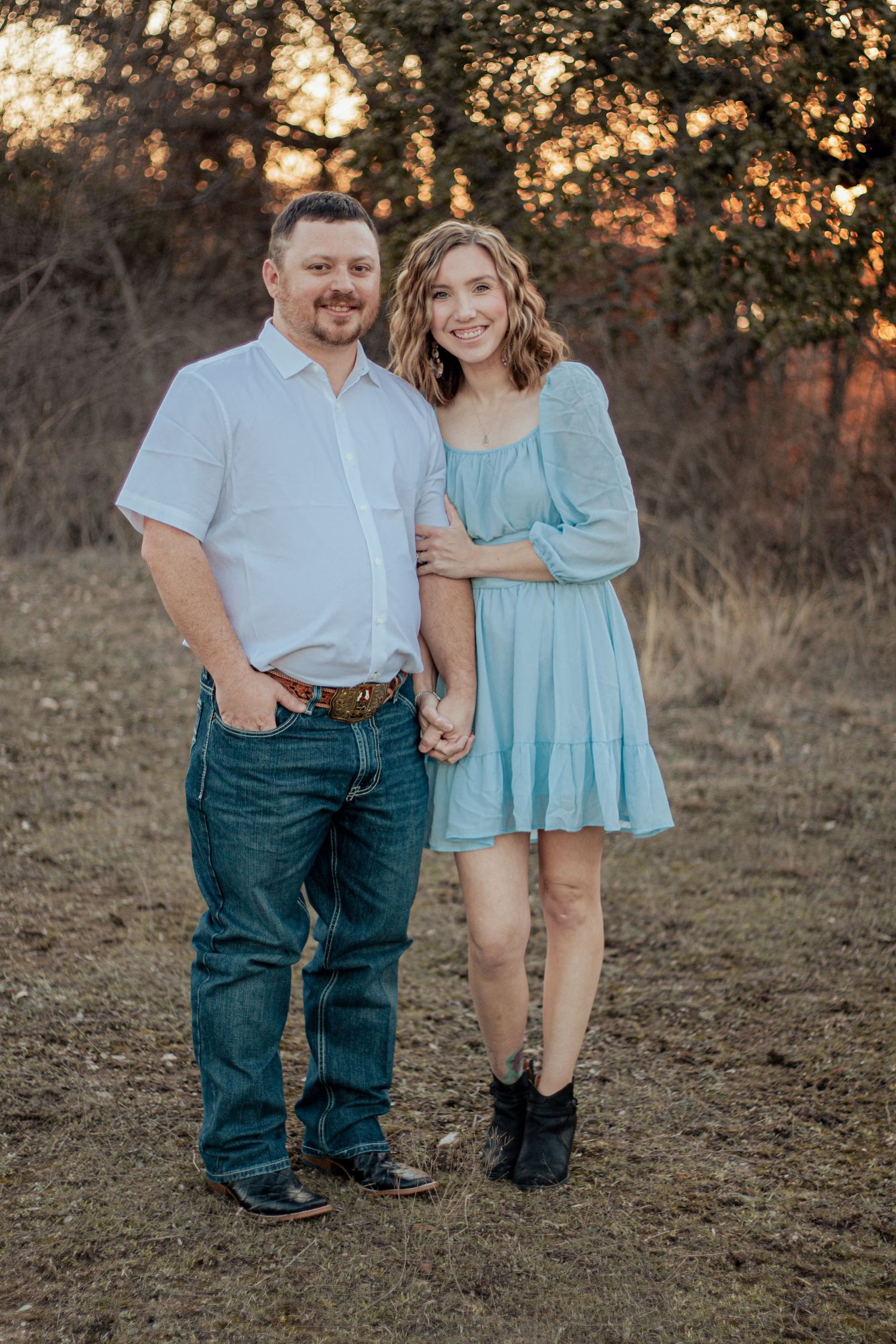 A couple poses outdoors at sunset, with the person in a light blue dress and another in a white shirt and jeans.
