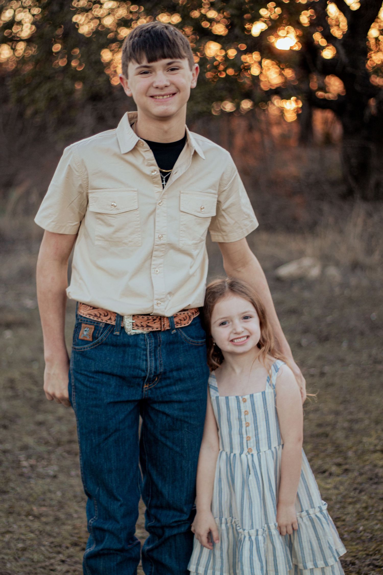 Two siblings stand together at sunset in outdoor portrait wearing casual western attire.