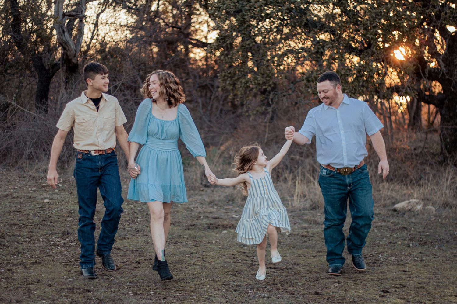 A group holds hands while walking together in a field at sunset, with sunlight filtering through trees in background.
