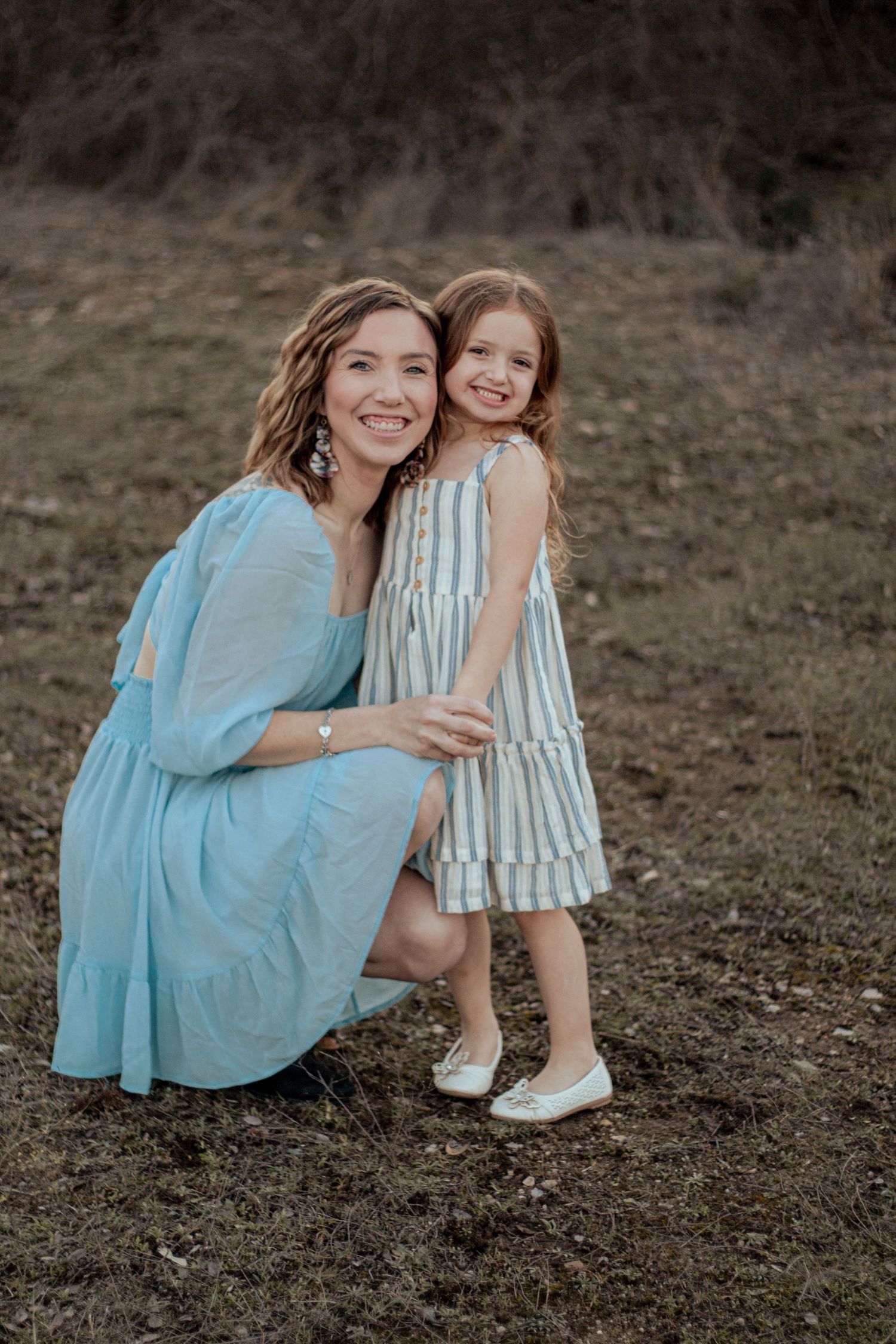 A woman in a blue dress and young girl in striped dress pose together outdoors at sunset.