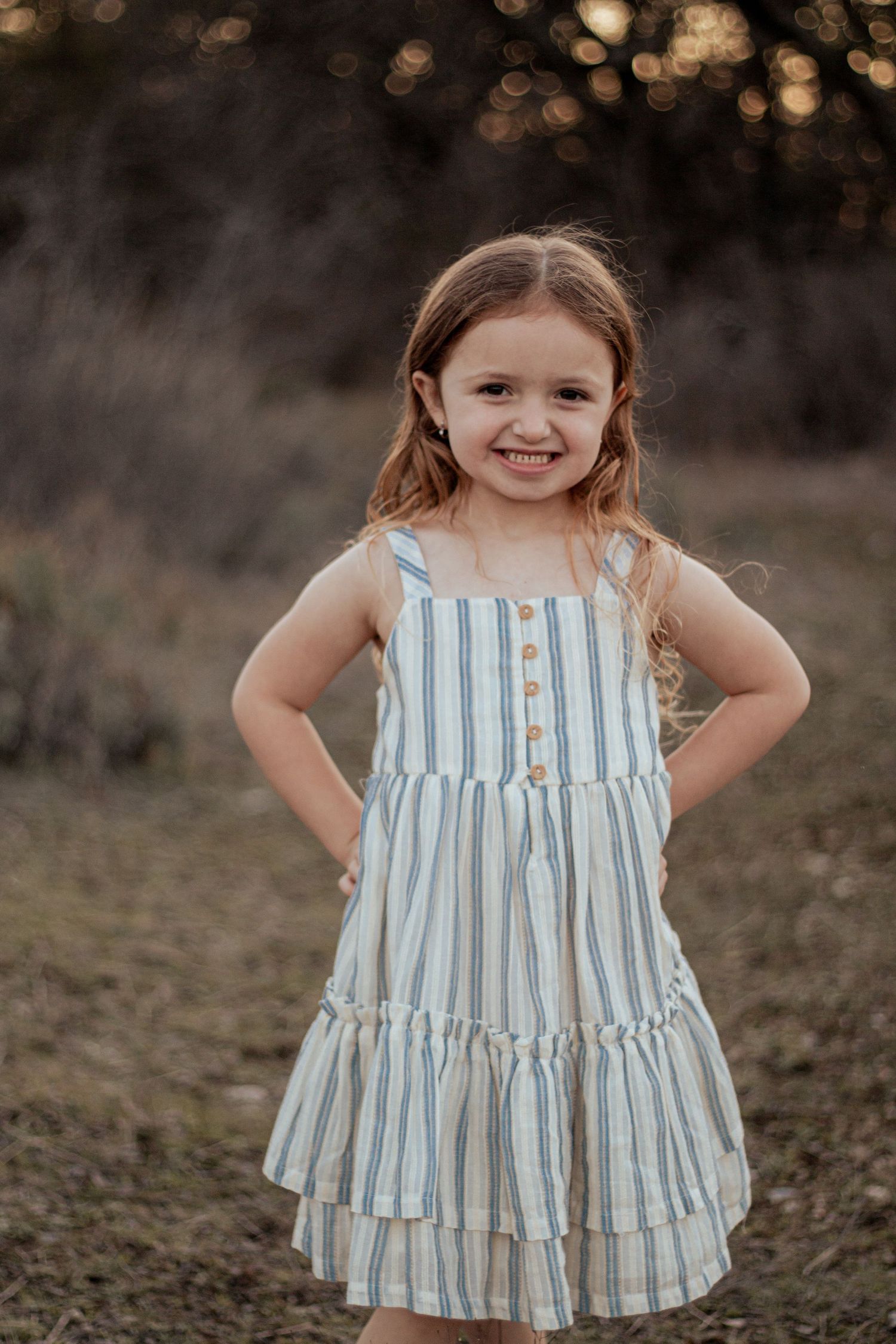 Young child poses outdoors in a blue and white striped tiered summer dress at sunset.