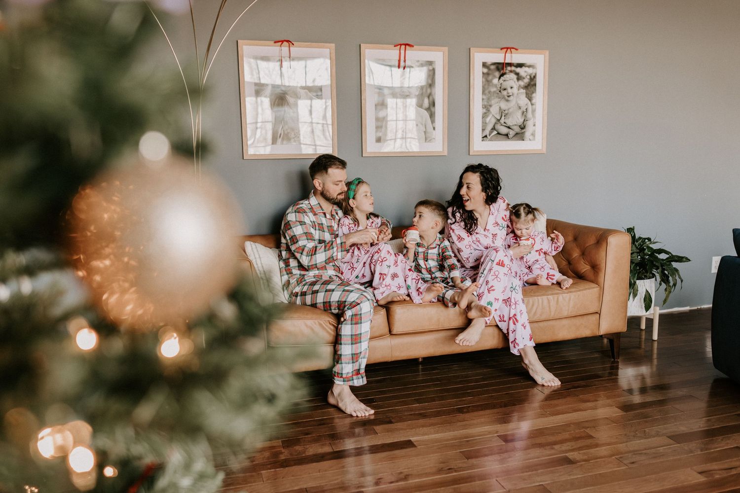 Two people in matching Christmas pajamas sit on a tan leather couch next to a decorated holiday tree.