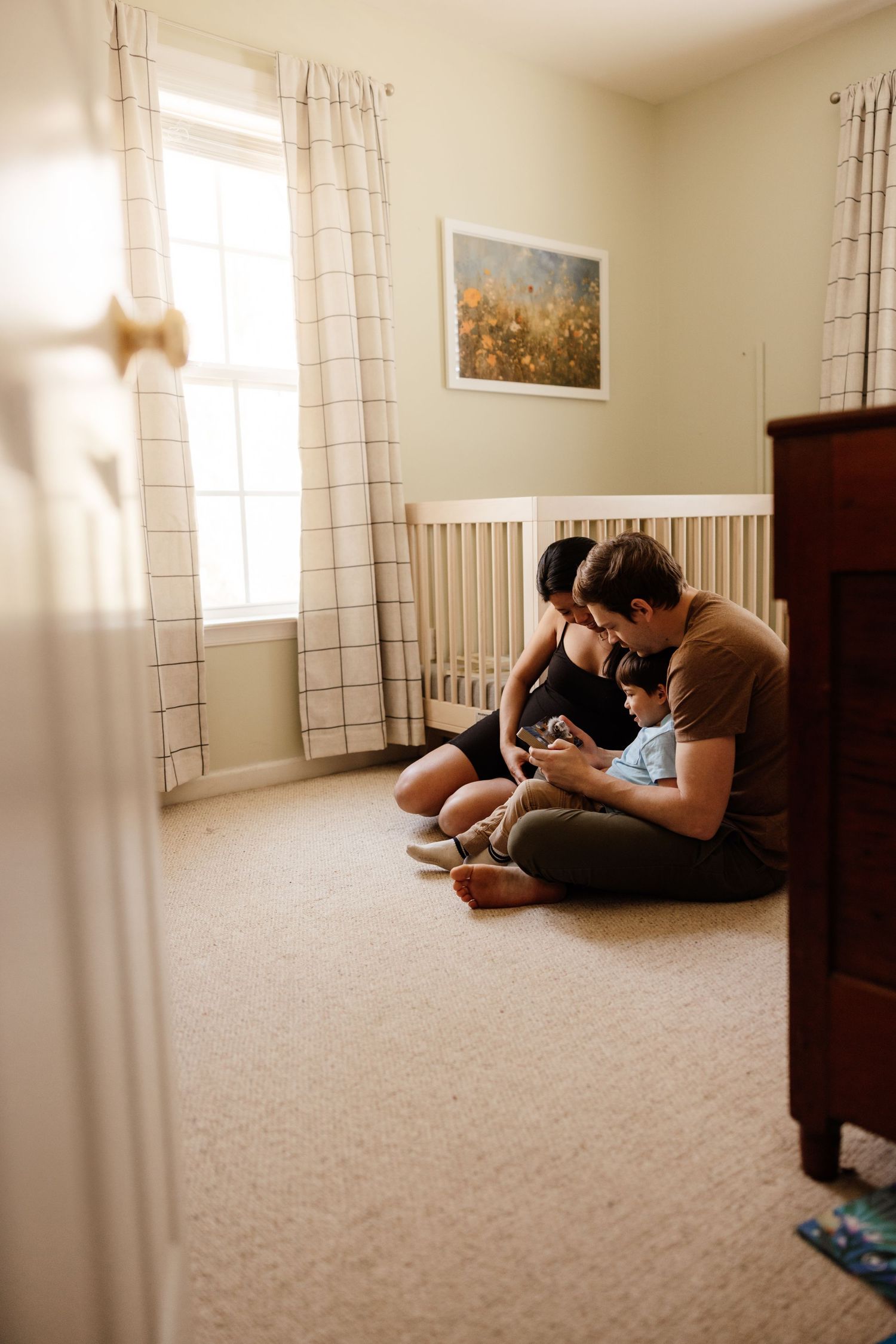 People sitting together on a carpeted floor of a sunny nursery room with white curtains and artwork on the wall.