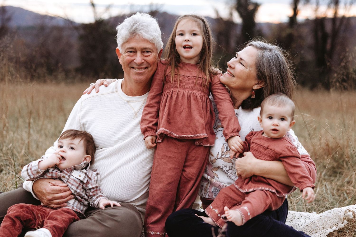 A family wearing coordinating rust-colored outfits sits together in a grassy field during sunset.