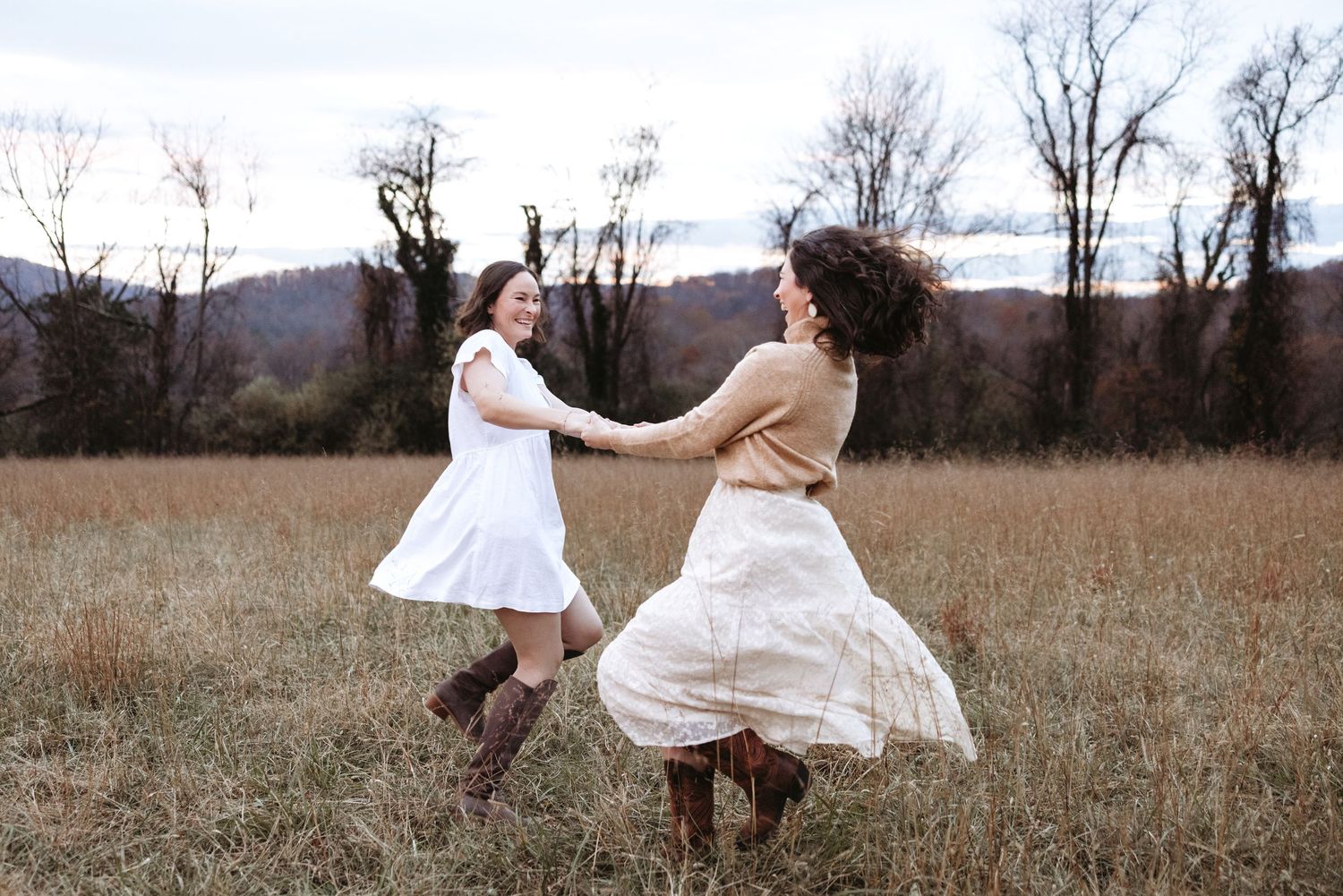 Two people in flowing white dresses dance and twirl together in a golden autumn field at sunset.