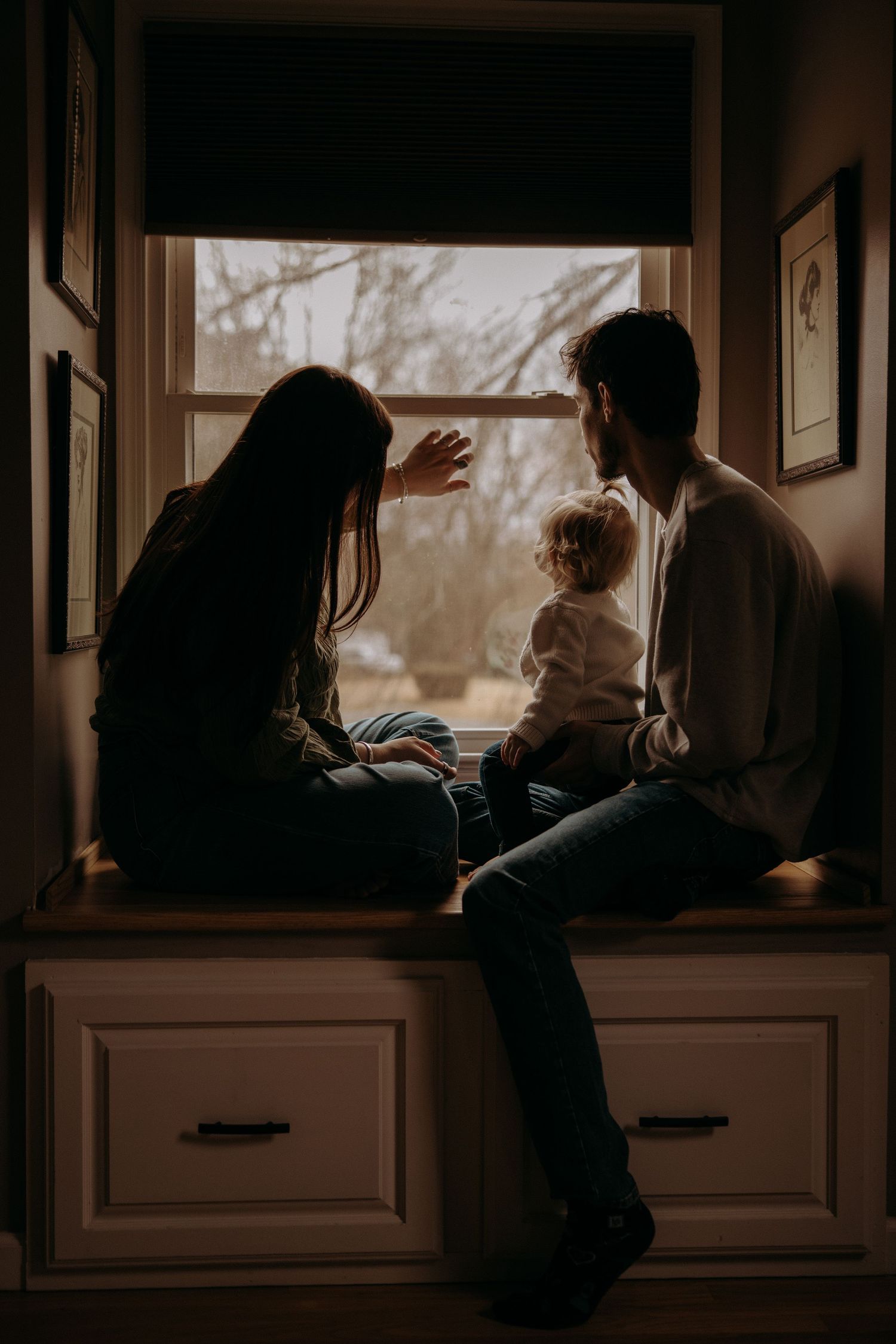 Family sits together in cozy window seat at home during golden hour creating intimate silhouette moment.