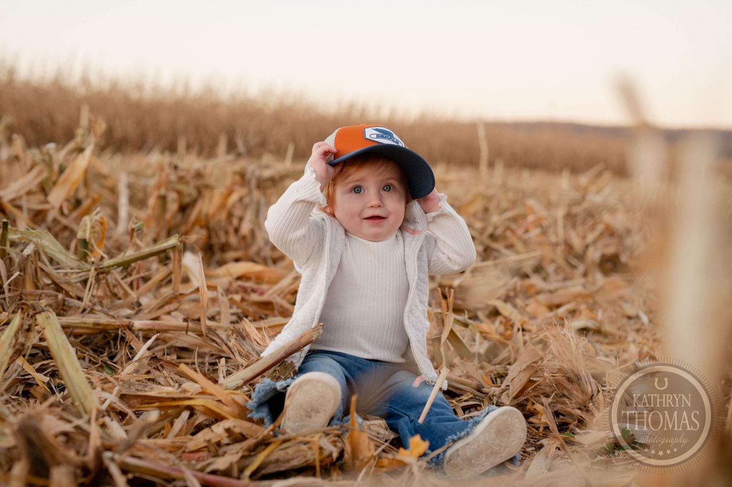 Young child wearing baseball cap sits among corn stalks in field.