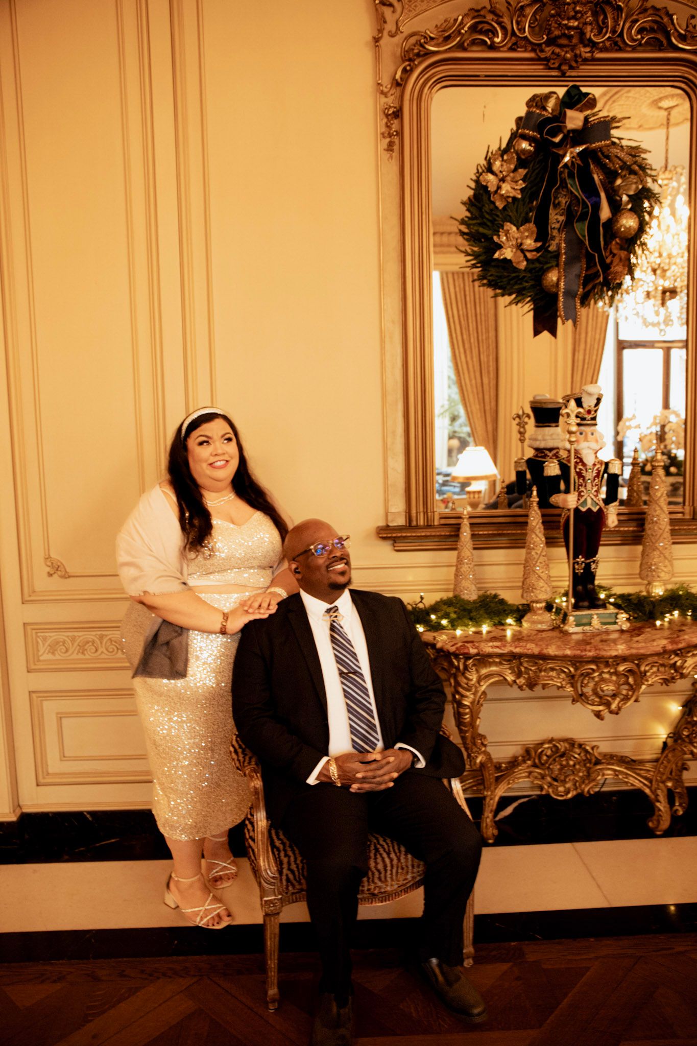 Elegant couple poses in formal attire next to ornate mirror with holiday wreath.