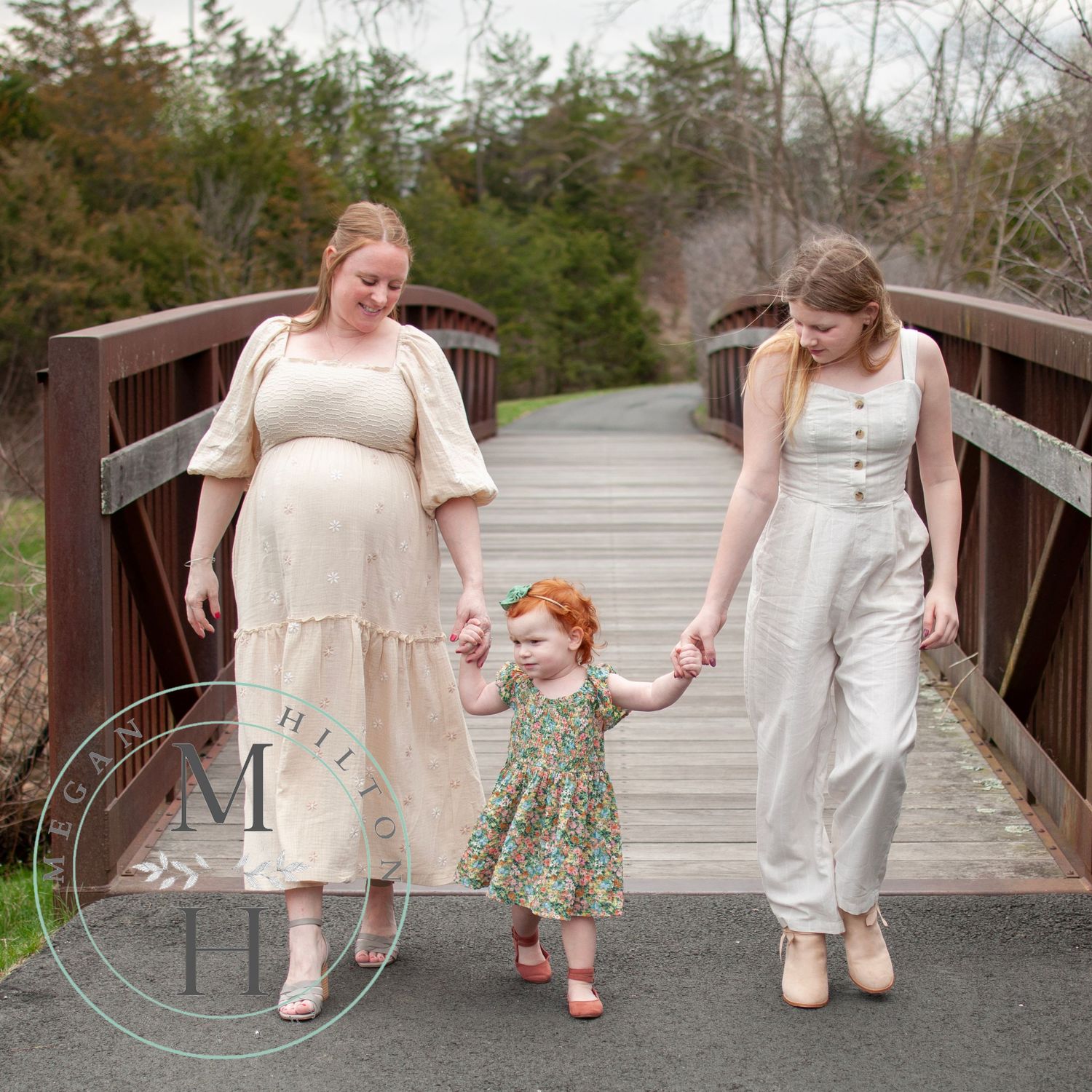 Photo of three girls walking hand in hand across a wooden bridge surrounded by trees on a spring day.