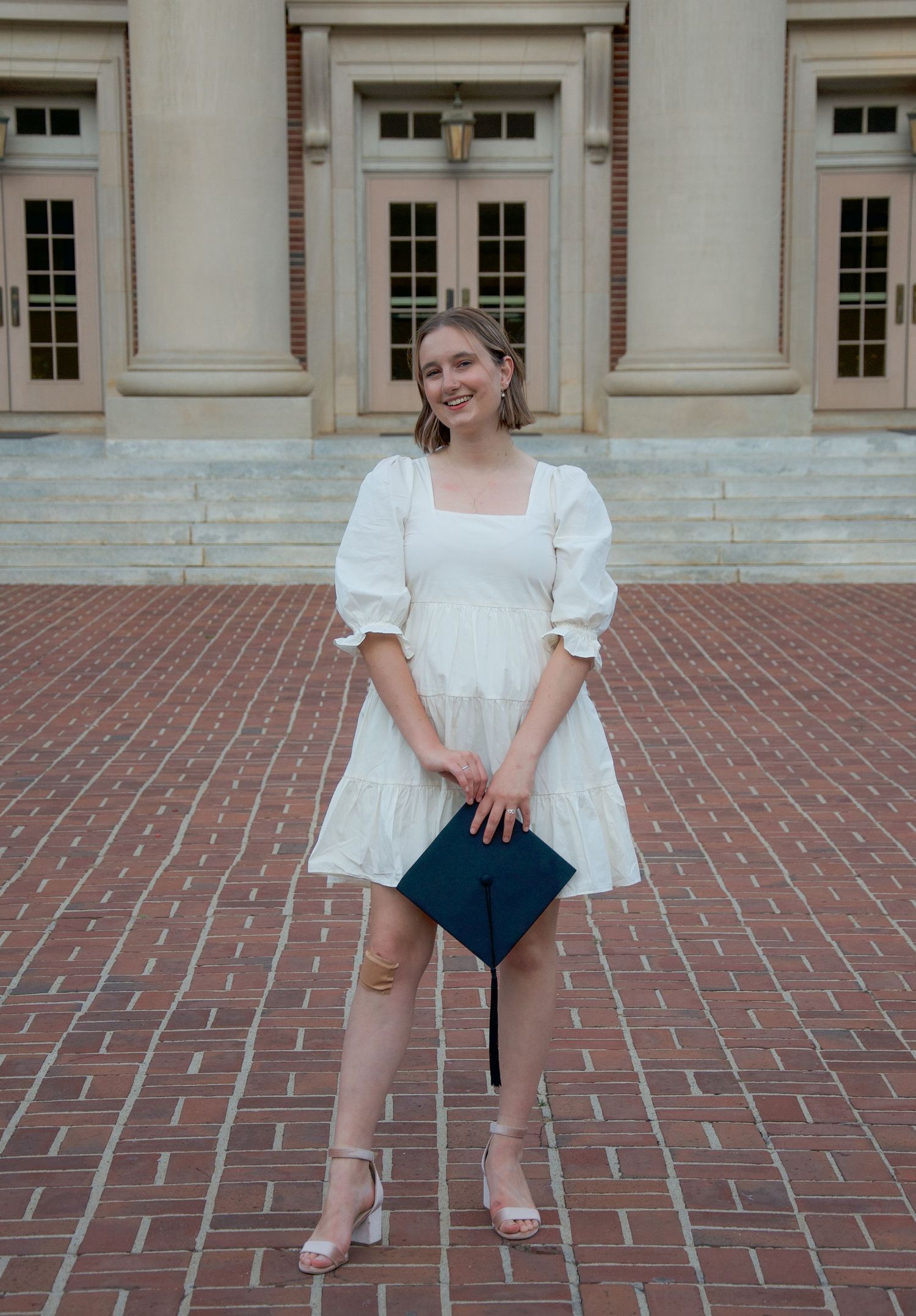 A young woman smiles for her college graduation portrait. She wears a white dress in front of her alma mater's main academic hall