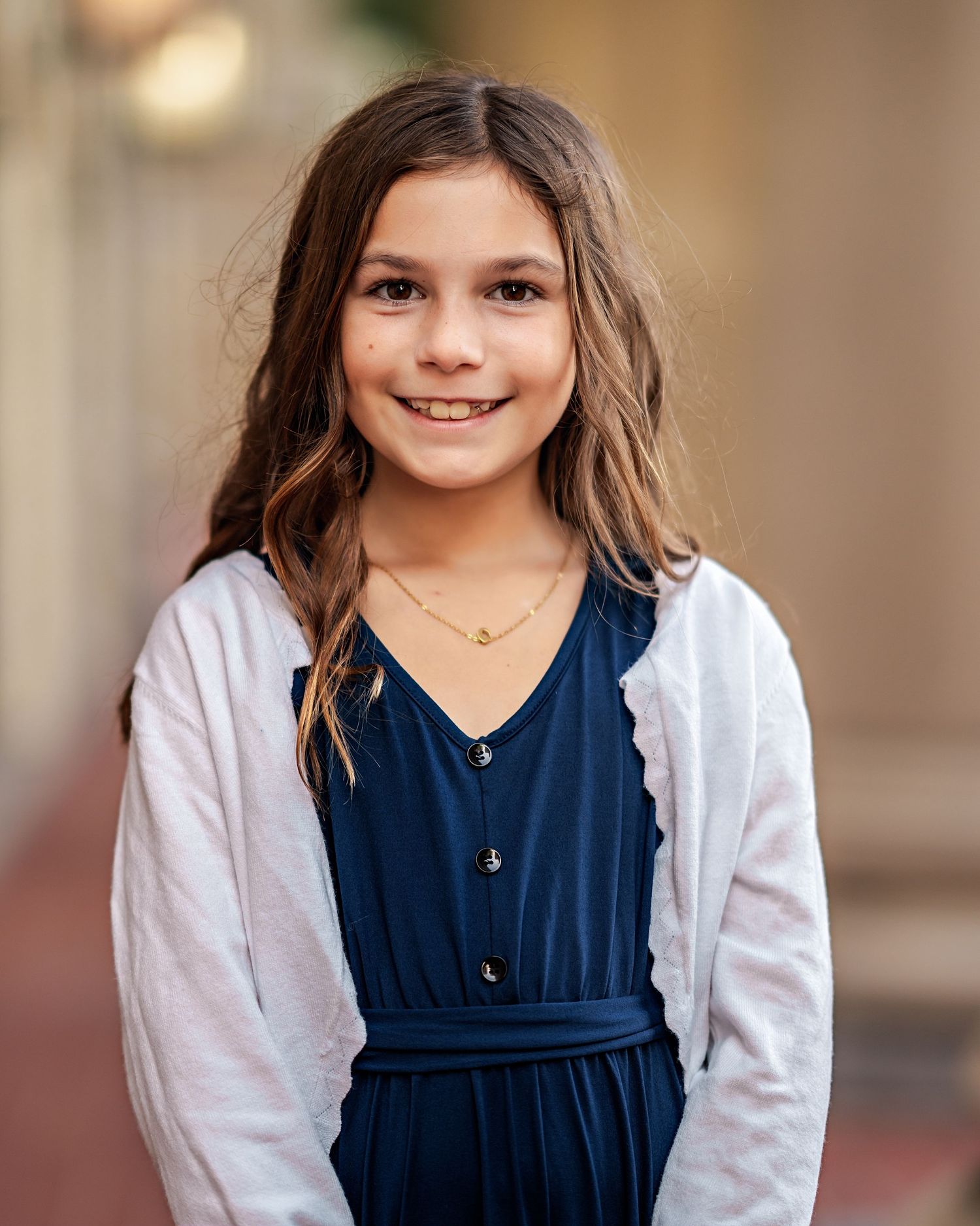 A young student in a navy blue dress with white cardigan and delicate necklace stands for a portrait photo.