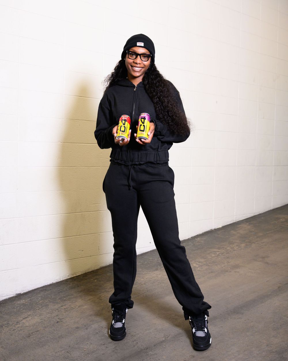 Person in black outfit holding energy drinks against white wall.