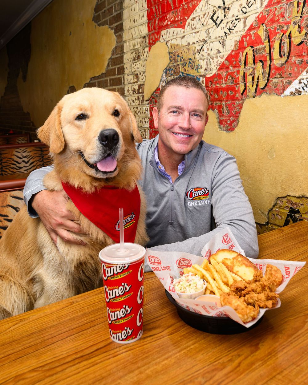 Man and golden retriever dog pose with Raising Cane's chicken fingers, fries, and drink at restaurant table.