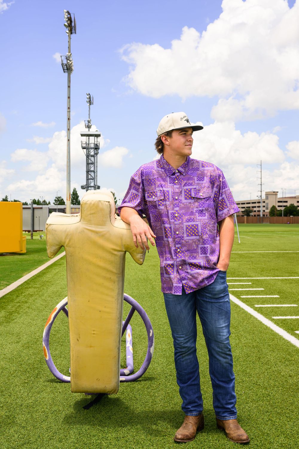 Young man in purple shirt and cap leans on football tackling dummy on a green field.