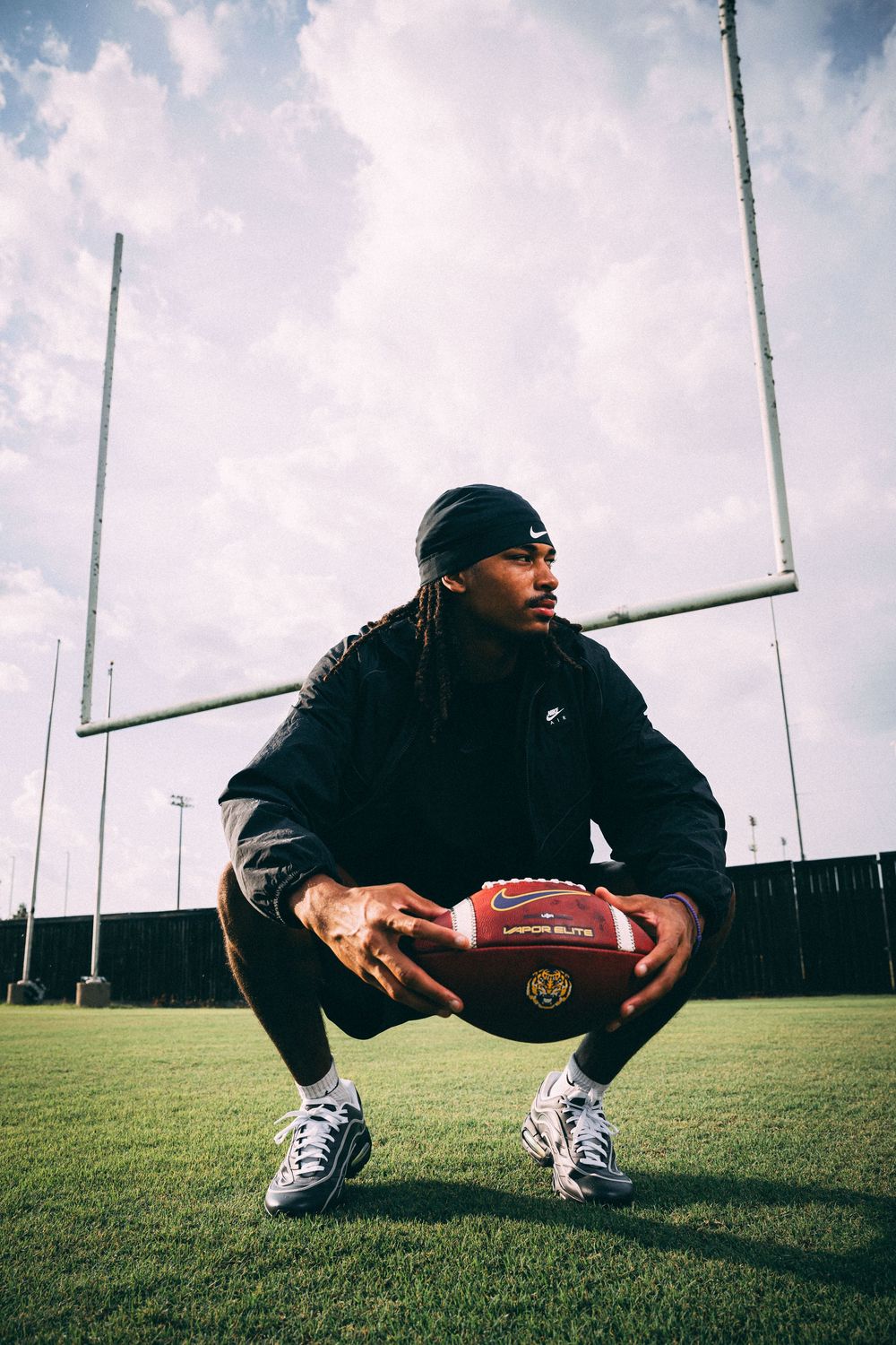 Football player in a black jacket and beanie crouches on a grass field holding a football in front of goalposts under a cloudy sky.
