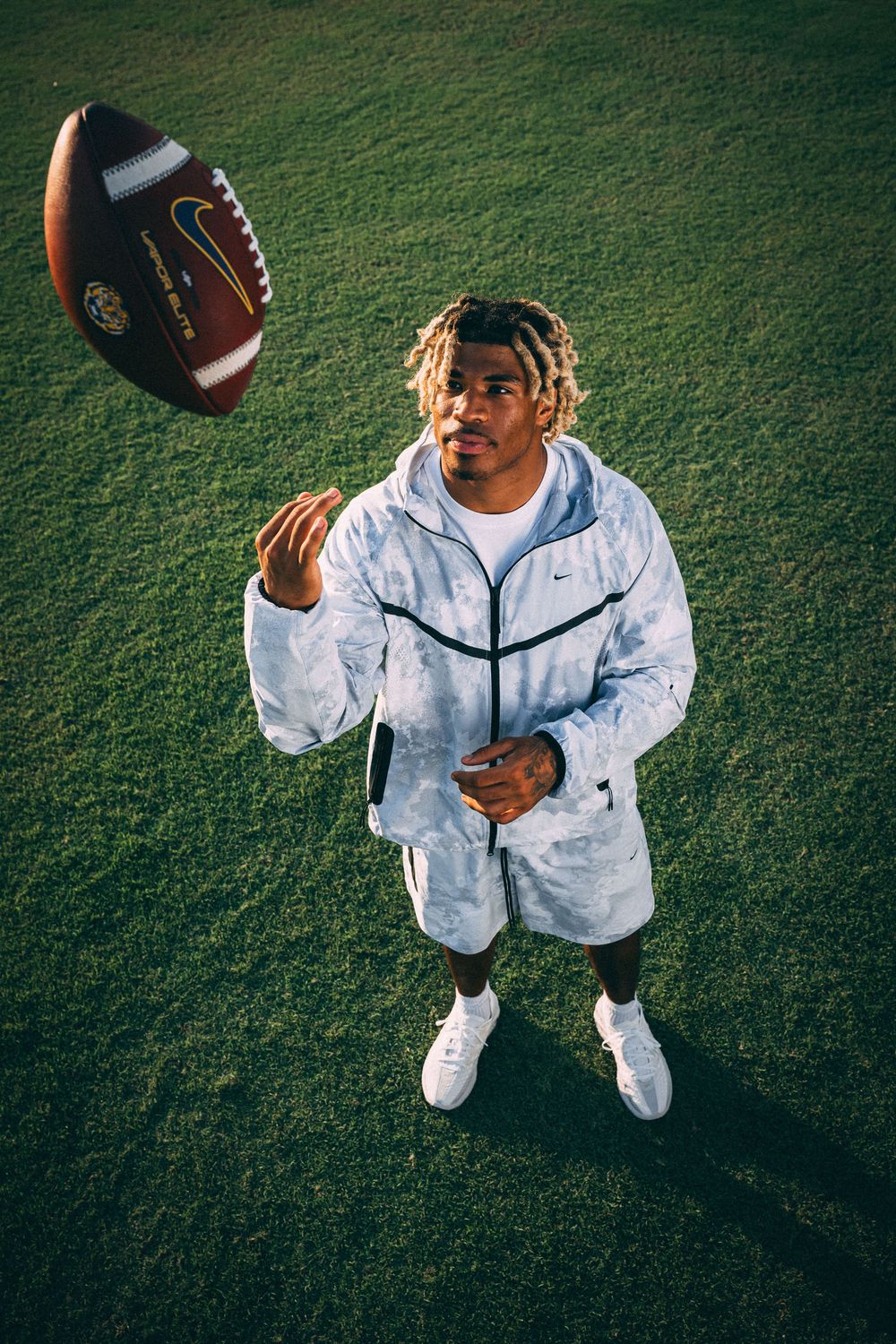 Athlete in a light gray Nike tracksuit tosses a football into the air while standing on a green grass field at sunset.