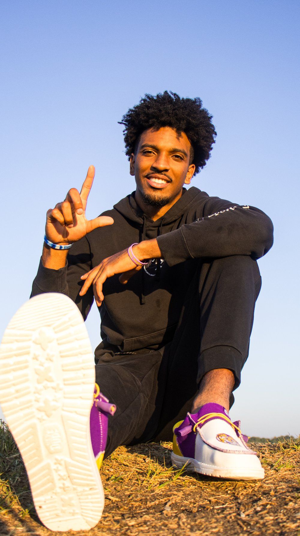 Athletic figure in black hoodie sitting outdoors making peace sign.