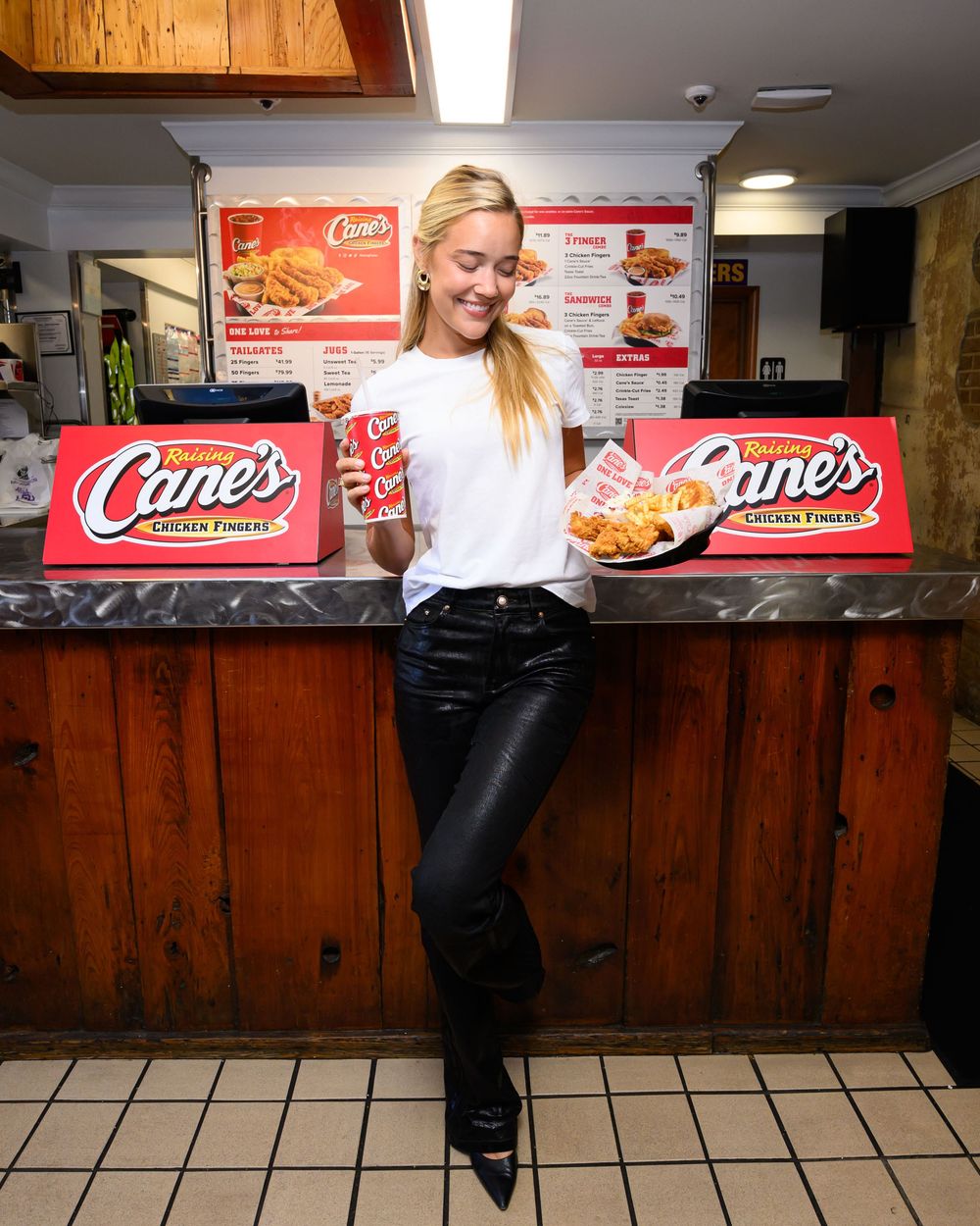Smiling woman in black leather pants leans on a counter at Raising Cane's, holding a cup and a tray of chicken fingers.