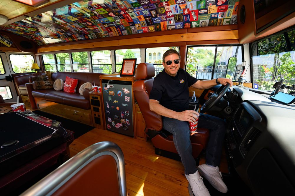 Smiling man in sunglasses sits in a leather driver's seat inside a colorful custom bus with a decorated ceiling, sofa, and wood floor.