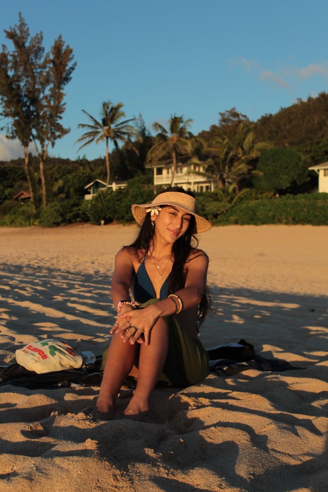 A beachgoer relaxes on sandy shores at sunset with palm trees and coastal homes in the background.