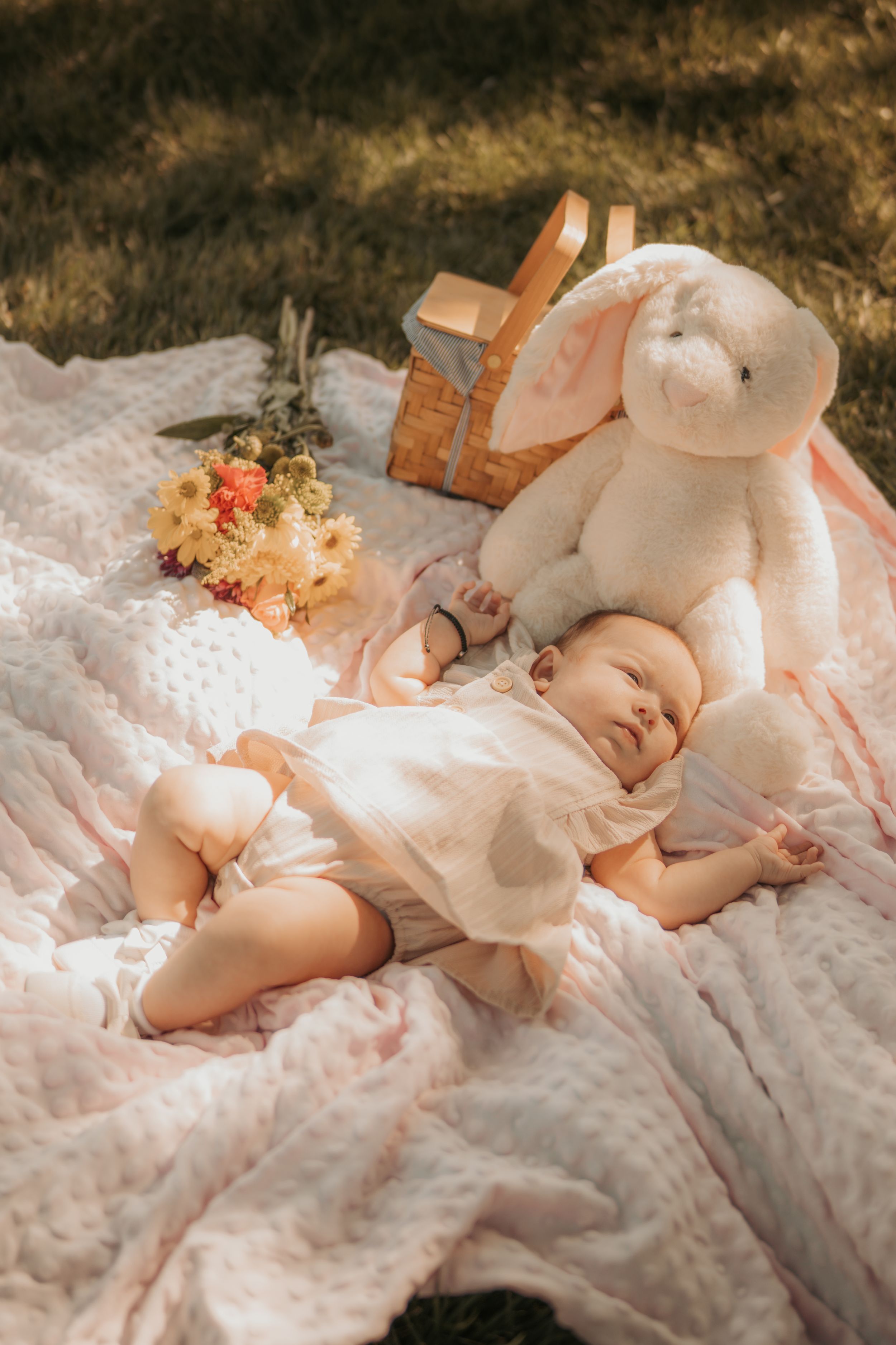 A baby rests peacefully on a white blanket outdoors with a plush bunny toy and picnic basket nearby.