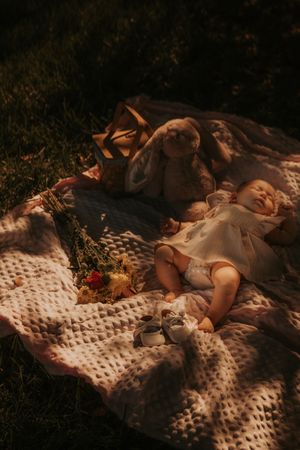 Baby laying on white knit blanket in dappled sunlight with stuffed toy bunny and autumn leaves during outdoor picnic.