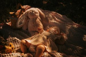 A child rests peacefully outdoors on a blanket next to a plush stuffed bunny toy in dappled sunlight.