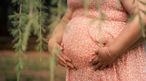 Someone in a peach-colored floral dress cradles their pregnant belly while standing outdoors near green foliage.