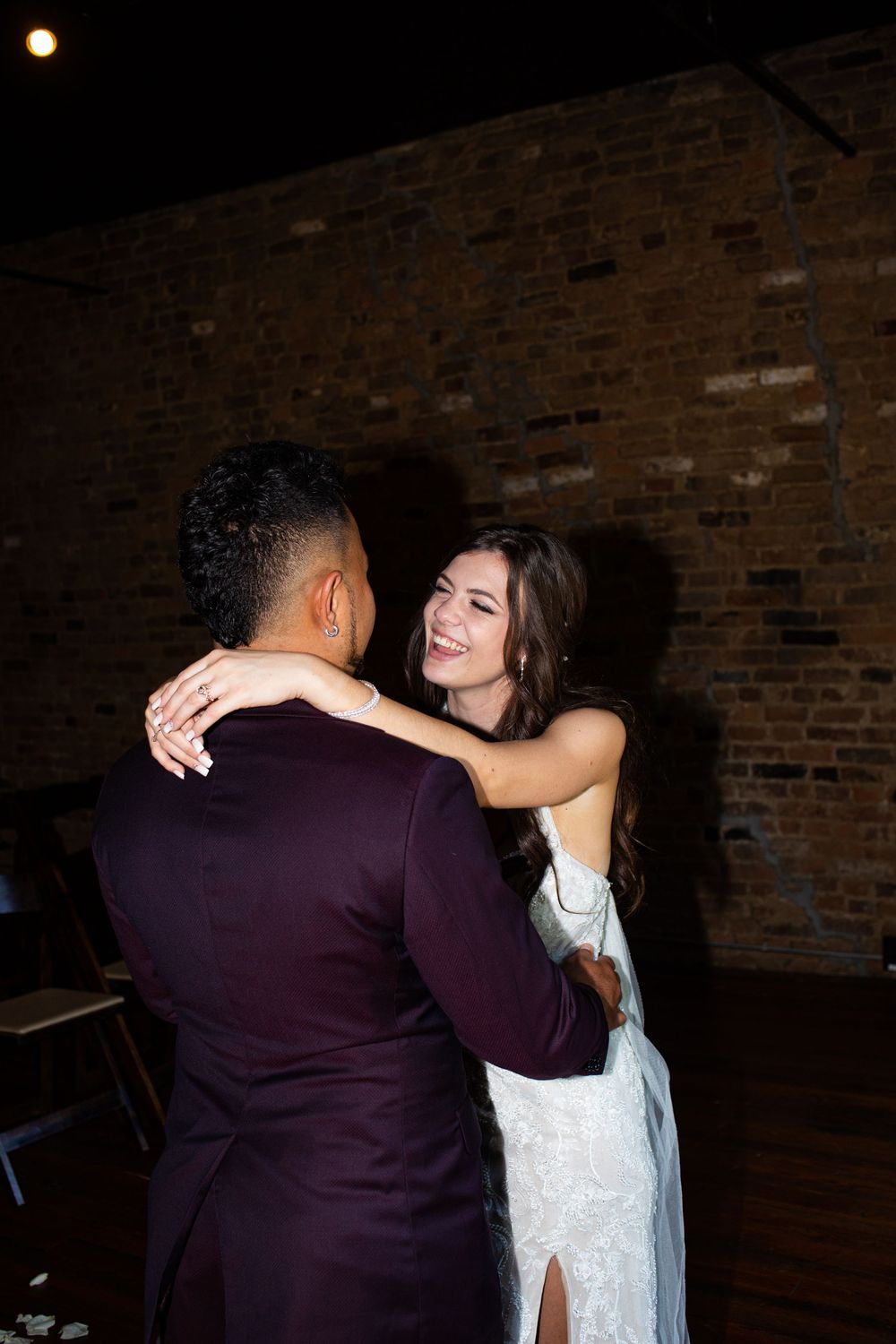 Bride in white gown smiling at groom in burgundy suit during first dance at rustic brick wall venue.