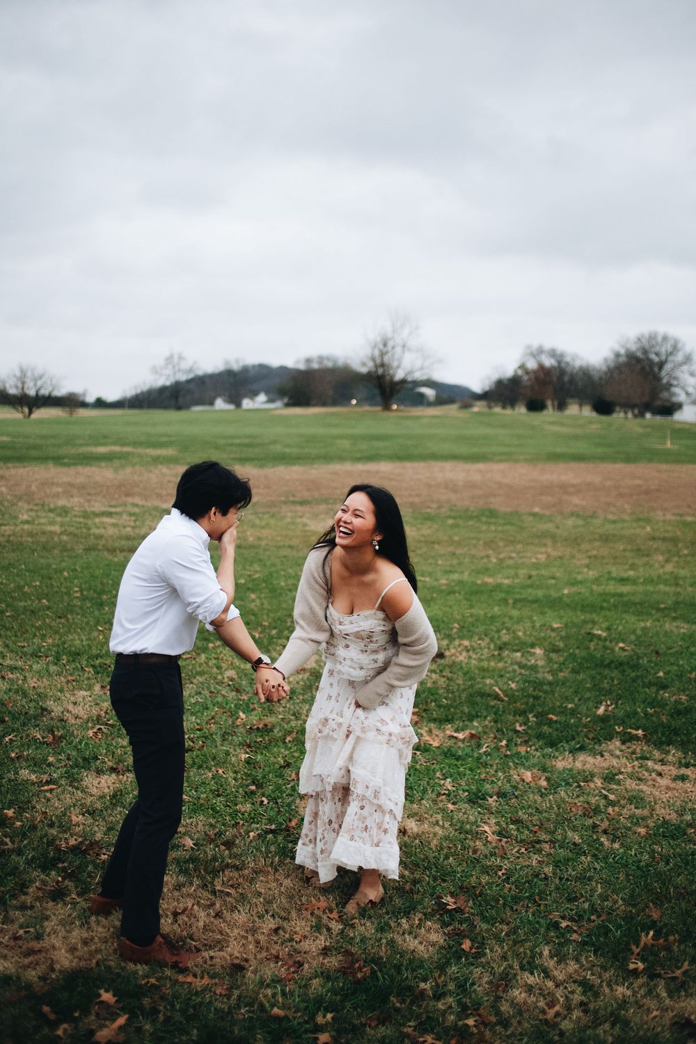 A couple in wedding attire playfully runs through an open field on a cloudy day.