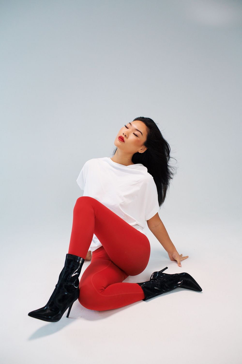 Woman in white tee, red leggings, and black heeled boots posing on floor against light gray background.
