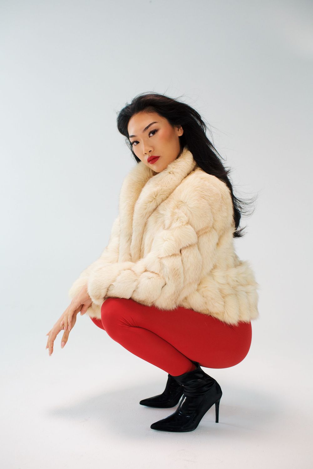 Woman in white fur coat, red pants, and black heels crouching against a light gray background.