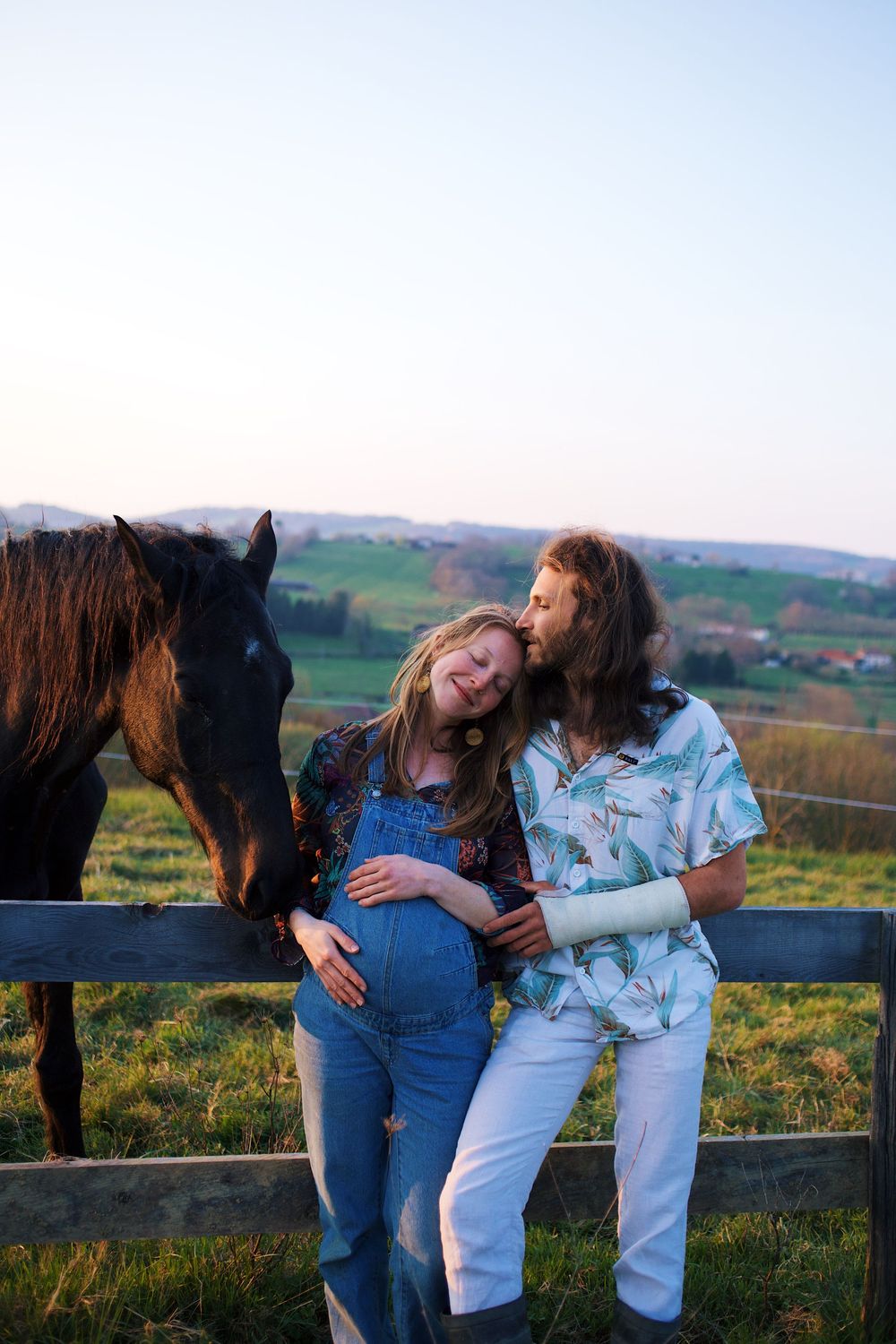 Pregnant couple embracing by a fence with a dark horse on a green hillside farm at sunset.