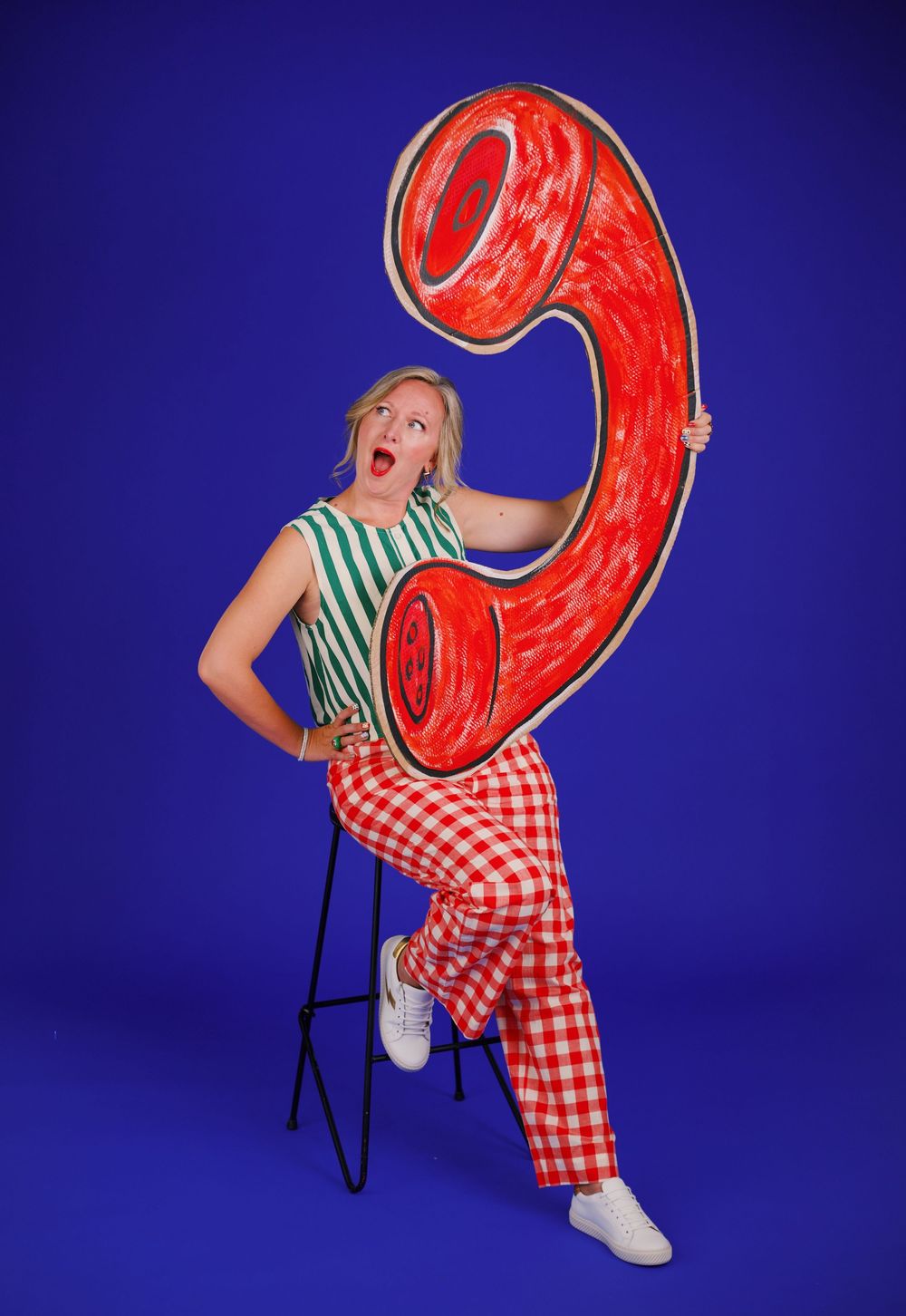 Woman in gingham pants poses on stool holding oversized red telephone prop against vibrant blue background.