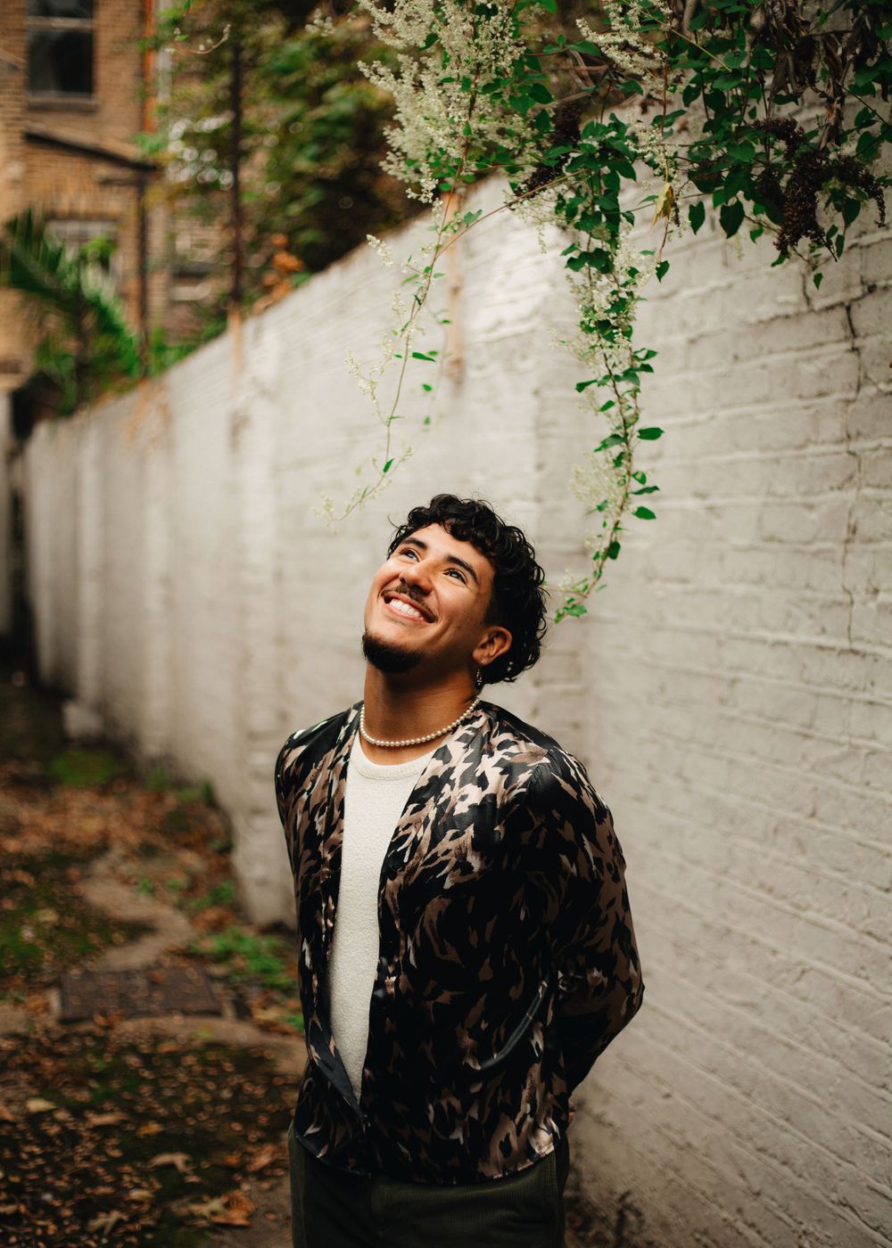 A person in a patterned jacket laughs joyfully against a white brick wall with hanging vines.