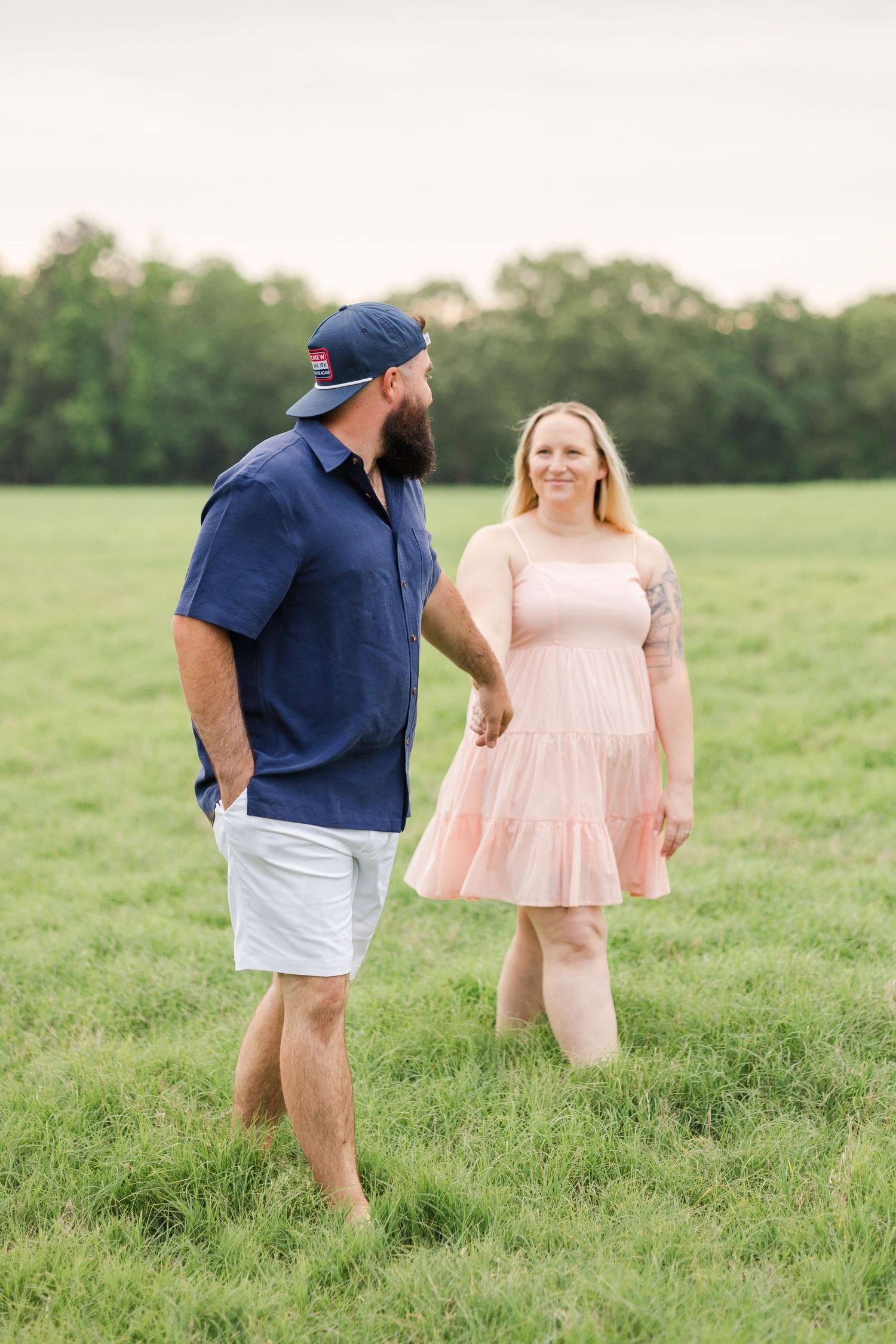 Couple in casual summer attire stands together in green field during sunset.