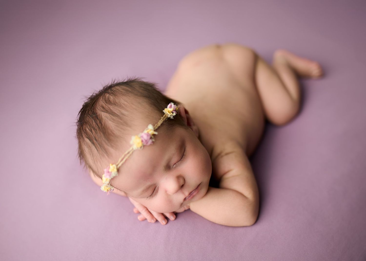 Peaceful sleeping newborn baby wearing delicate floral headband rests on soft purple backdrop during professional photo session.