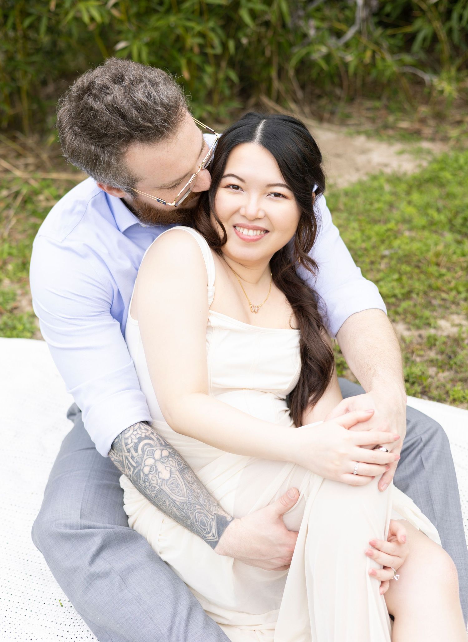 A couple sits intimately together outdoors on a blanket, sharing a tender moment against a natural backdrop.