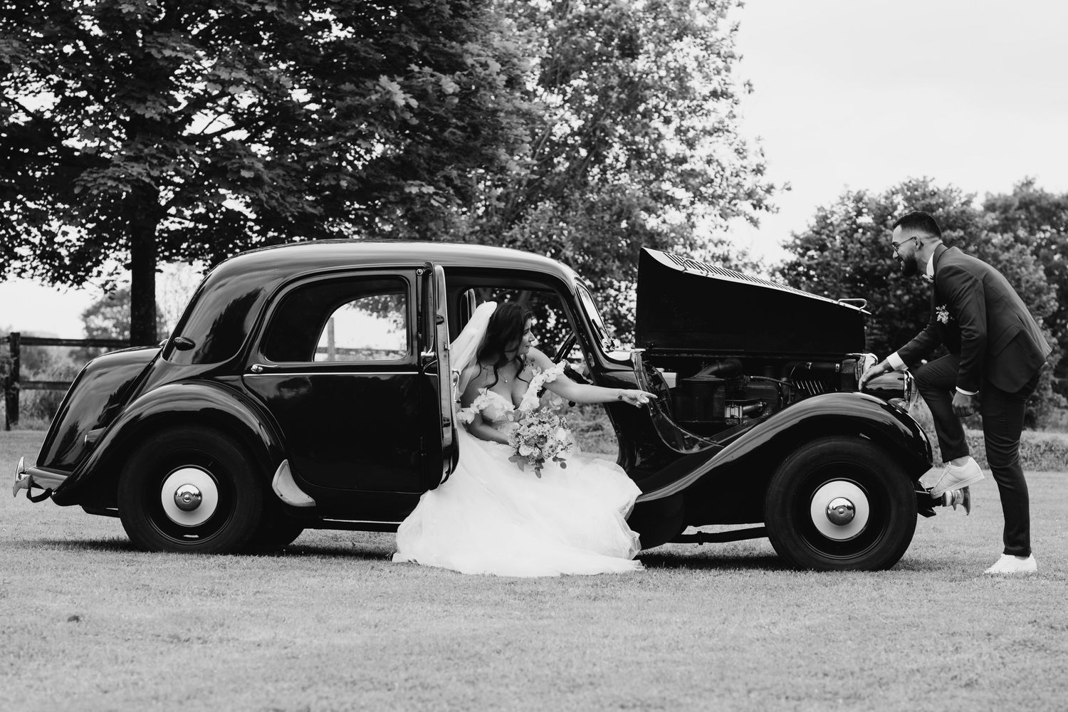 Black and white photo of a bride posing beside a vintage classic car with groom.