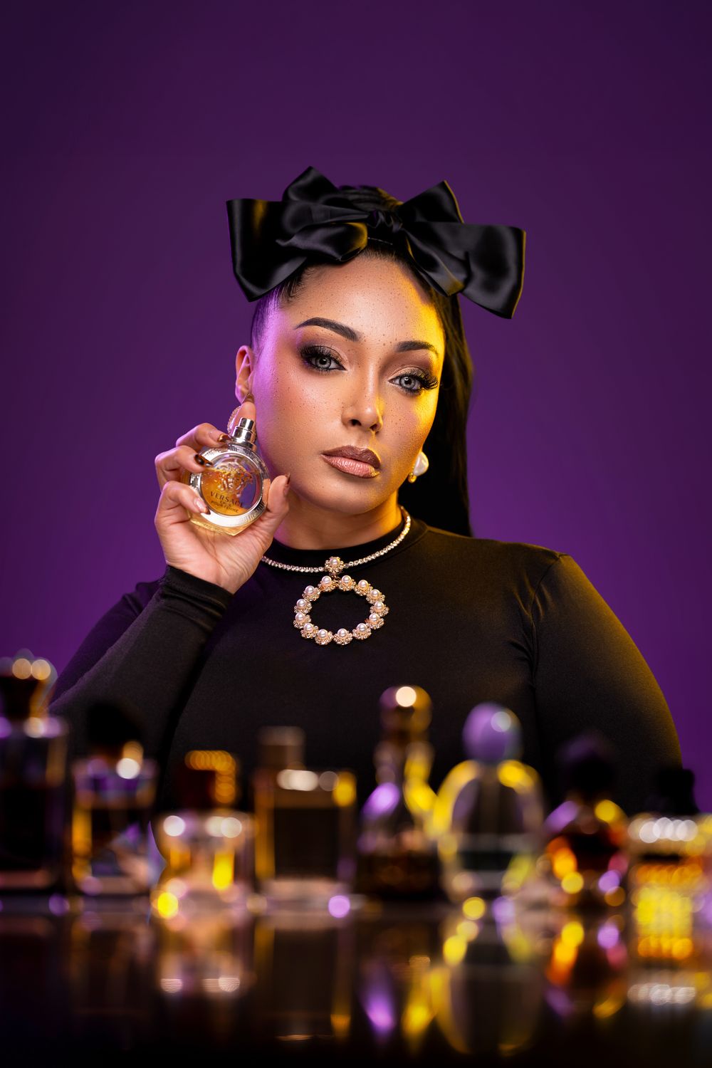 Glamorous person in black attire with bow headpiece holding perfume bottle against purple backdrop with blurred bottles.